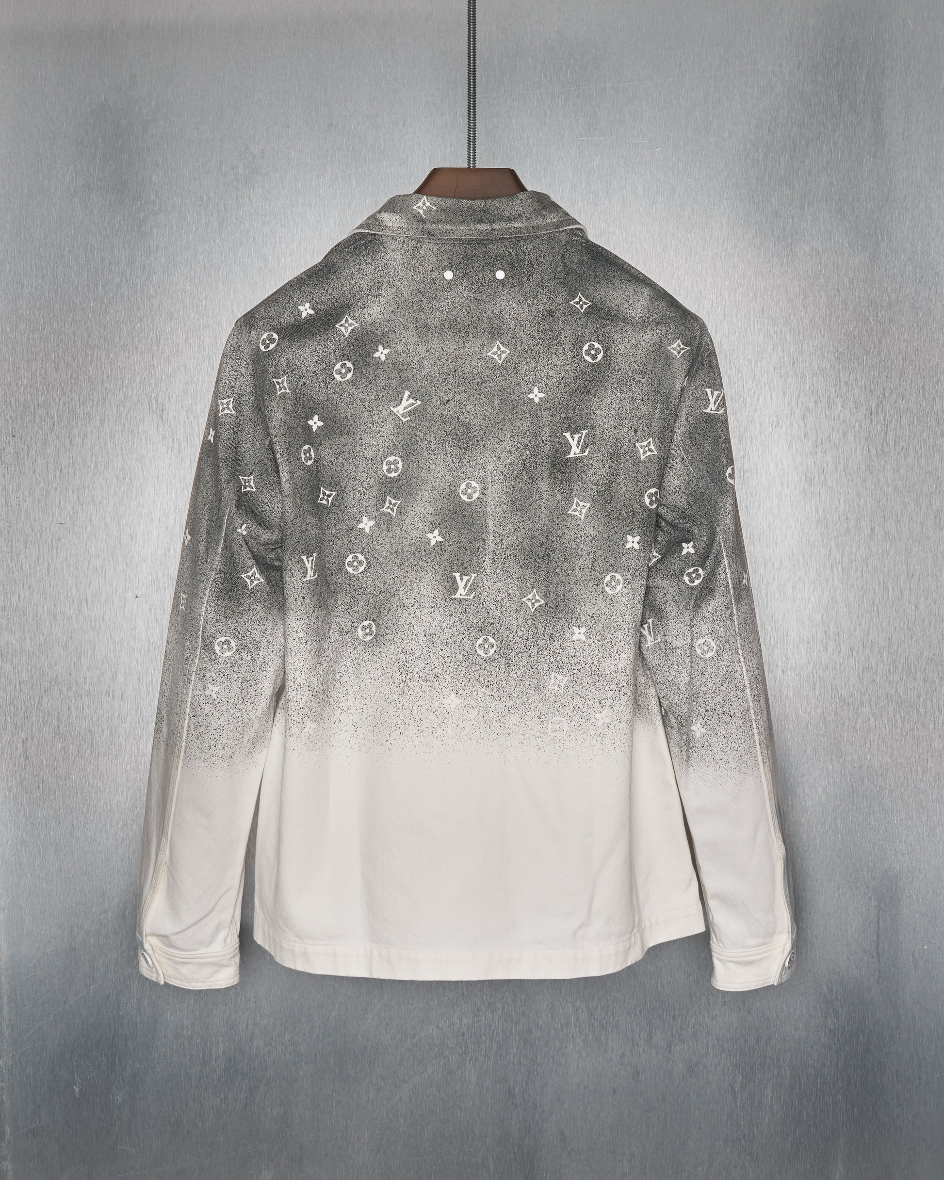 Reverse of a white Louis Vuitton jacket with a gradient black spray effect on top, featuring small, repeated white logos. It hangs on a wooden hanger against a steel backdrop.