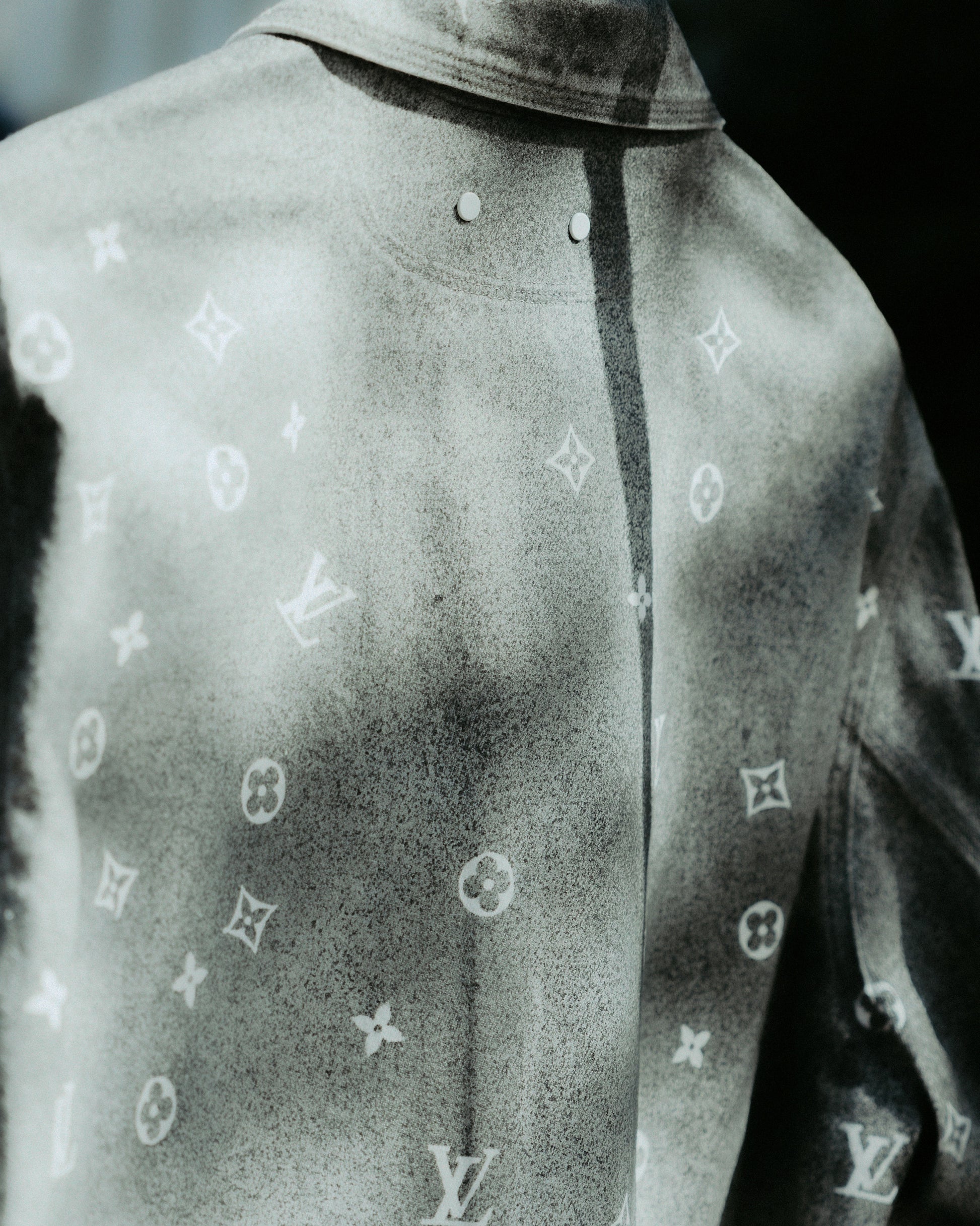 Close-up of a grey Louis Vuitton jacket featuring a subtle, ornate pattern with a glossy finish. The pattern repeats logos and floral motifs
