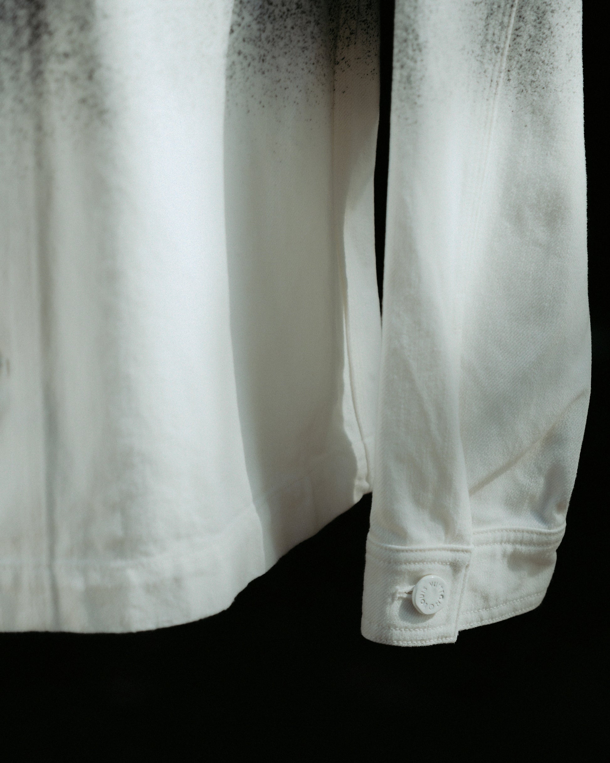 Close-up of a white Louis Vuitton shirt sleeve with a button on the cuff. The fabric has subtle black speckles at the top, creating a soft gradient. The mood is calm and minimalist.