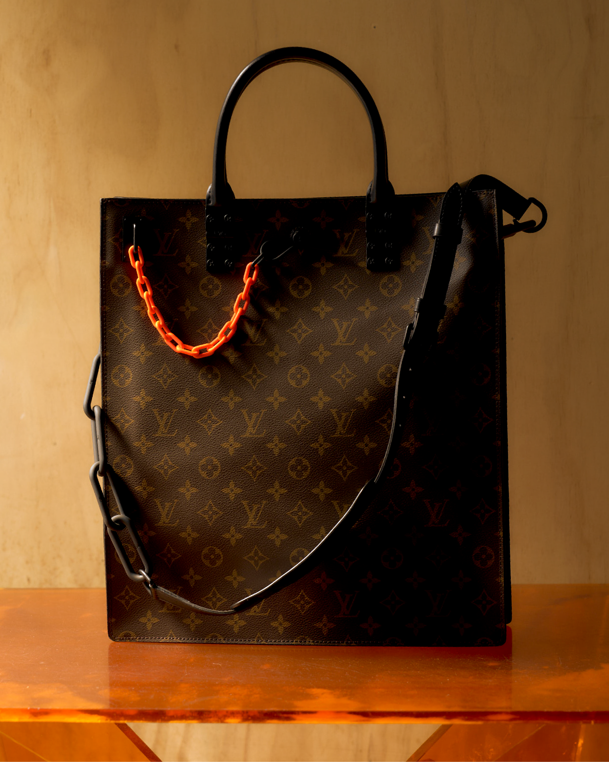 Louis Vuitton monogram tote bag with orange chain, matte black leather detailing, and a visible logo on the fabric.