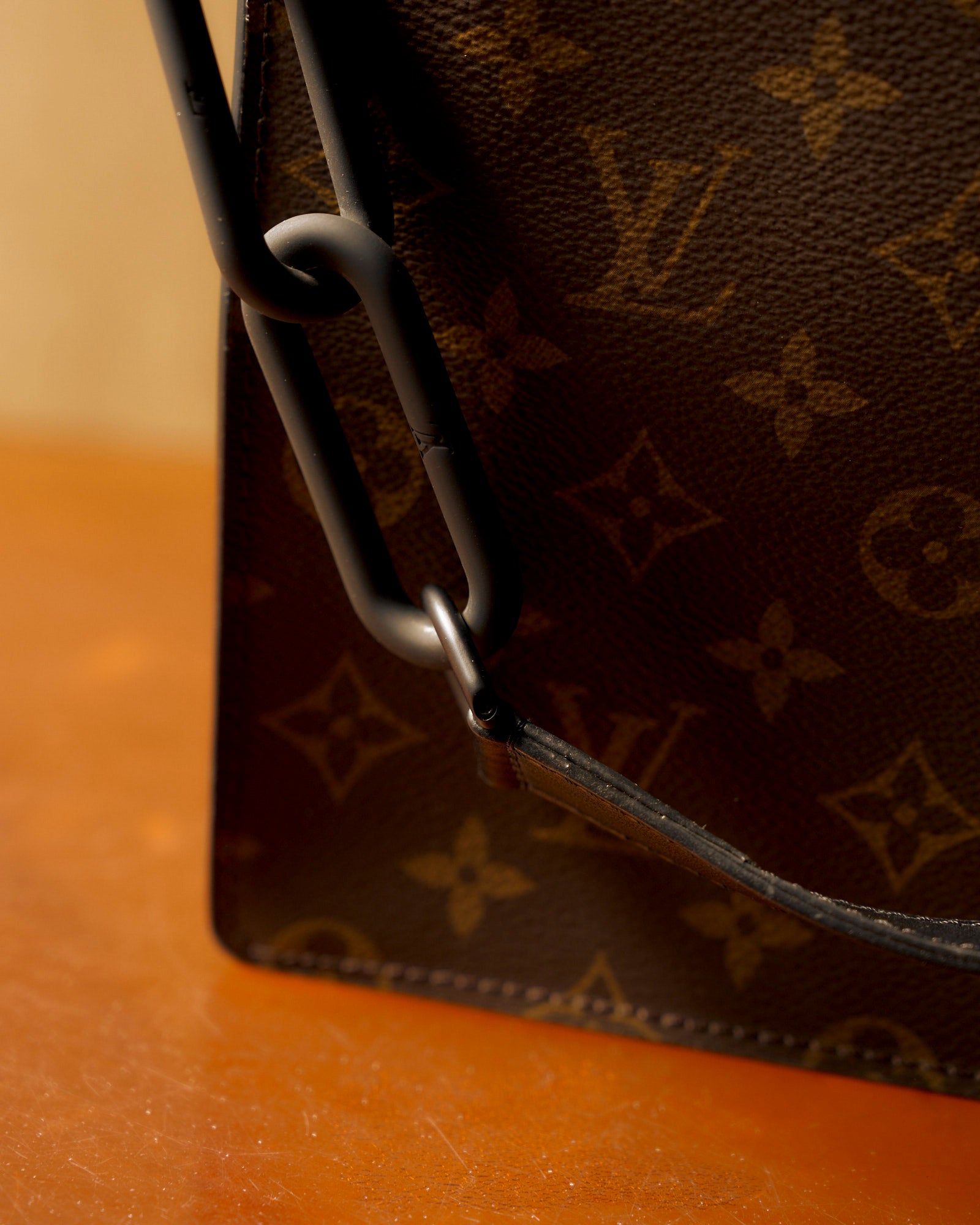 Close-up of a Louis Vuitton handbag corner with a leather monogram pattern and a large black metal chain handle against a warm, light brown surface.