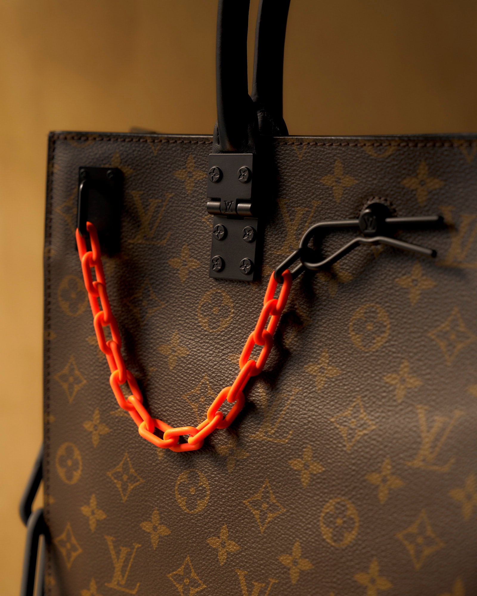 Close-up of a Louis Vuitton handbag featuring a brown monogram pattern with black handles. A bold orange chain adds a pop of colour