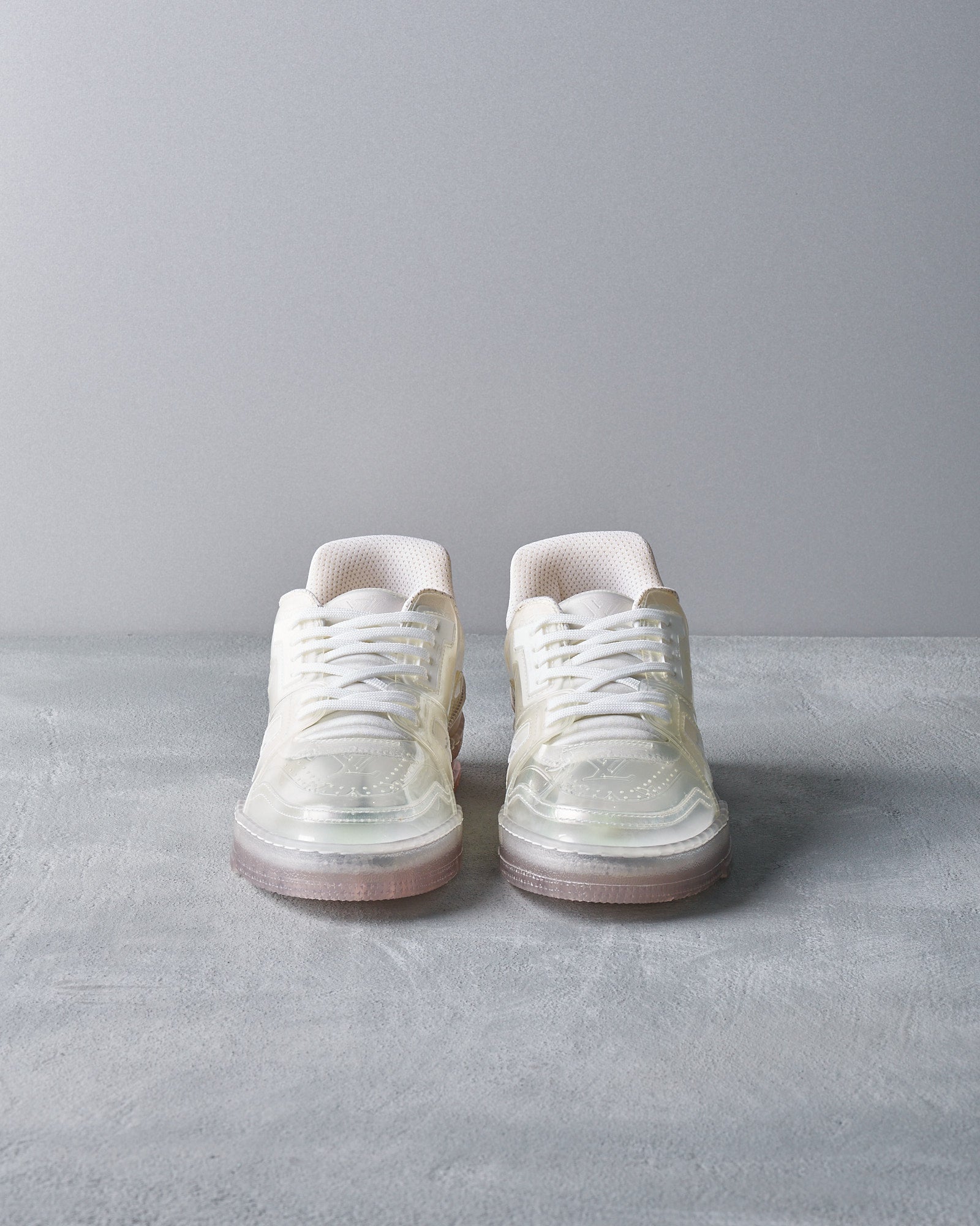 Stylish white Louis Vuitton sneakers on a grey textured floor. The shoes have glossy semi-transparent overlays and pinkish translucent soles. Modern and sleek design.