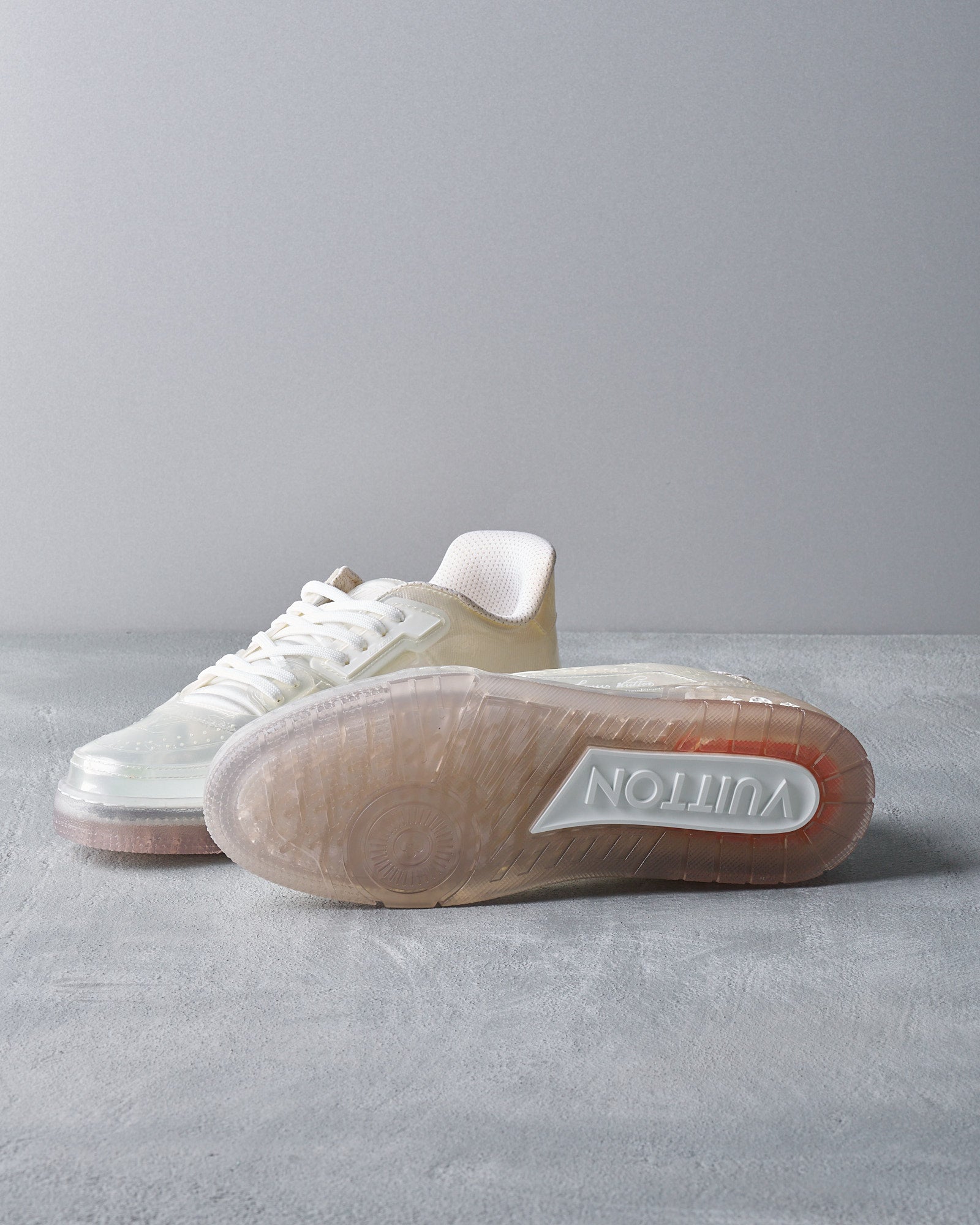 A pair of stylish beige and white sneakers on a gray surface. One shoe lies flat while the other is on its side, showing a translucent sole with "Vuitton" embossed in white. The tone is modern and trendy.