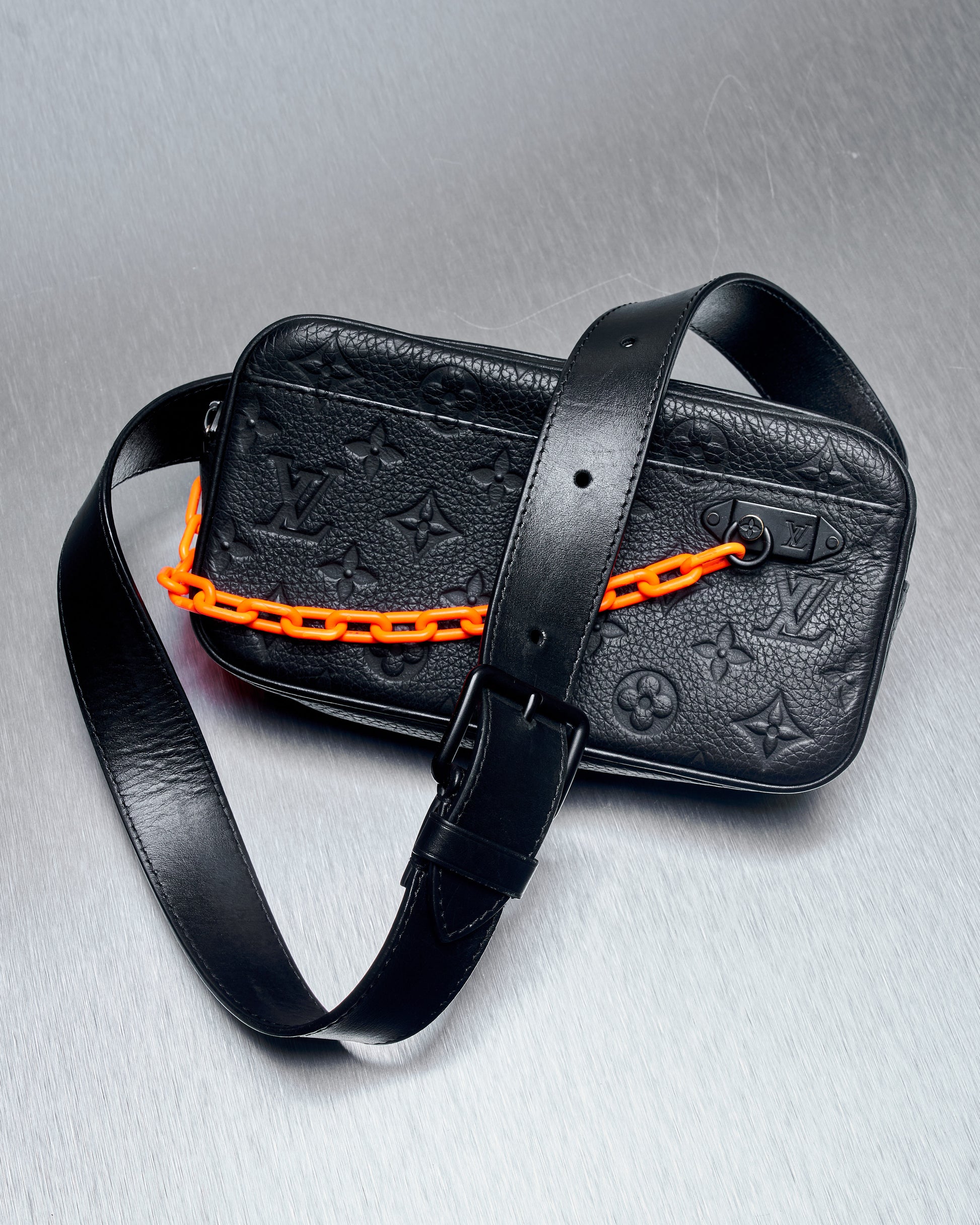A black leather Louis Vuitton crossbody bag with embossed monogram pattern, featuring a bold orange chain and matching strap, lying on a textured silver surface.
