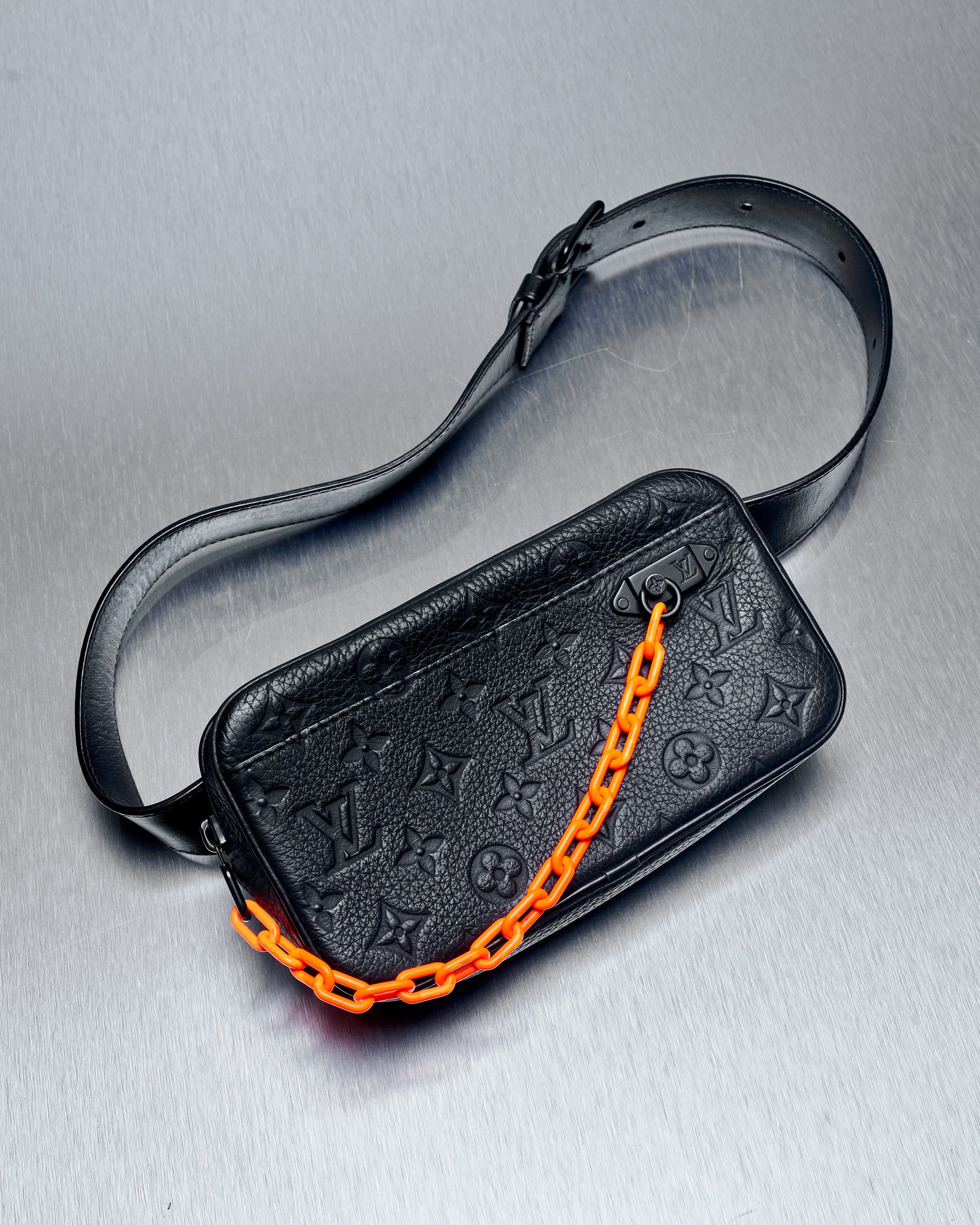 A black Louis Vuitton belt bag with an orange Solar Ray chain, featuring a front flap pocket and tonal metal hardware.
