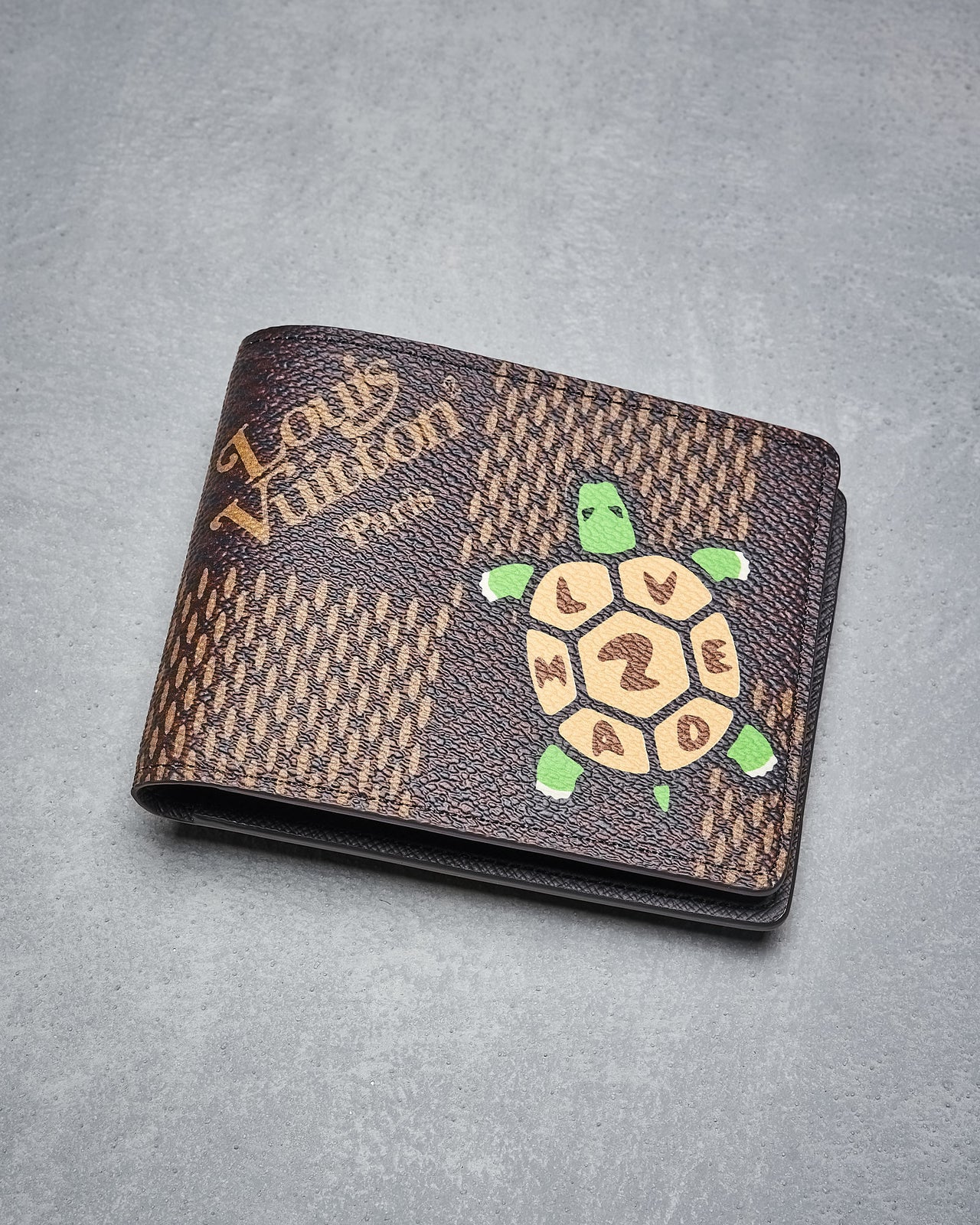 The Louis Vuitton x Nigo wallet damier ebene giant brown in coated canvas features a printed green turtle with "LIVE, HARD," and the number 2 on its shell, shown on a gray textured surface.