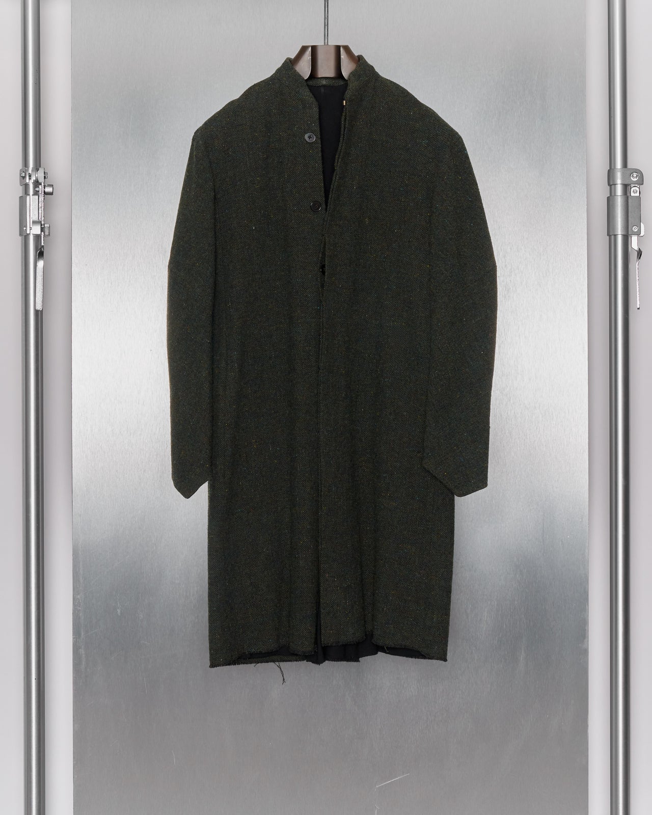 A long, mottled green wool coat from M.A+ hanging on a coat rack, featuring a collarless design, hidden button front, and distressed hem detailing.