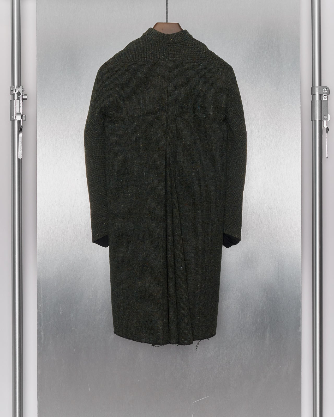 A long, mottled green wool coat from M.A+ hanging on a coat rack, featuring a collarless design, hidden button front, and distressed hem detailing.