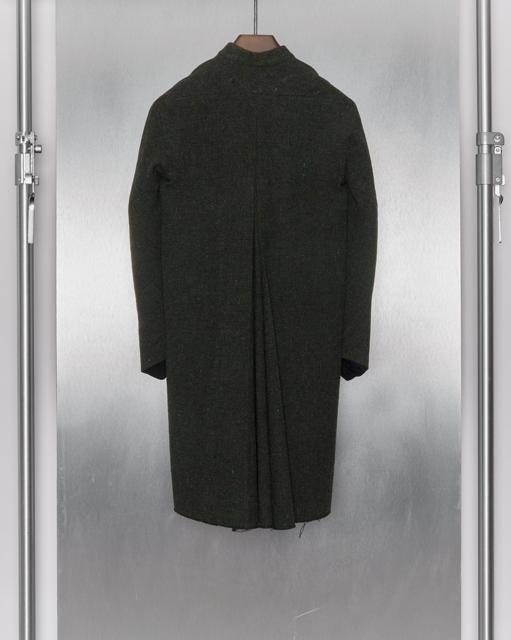 Reverse of a long, mottled green wool coat from M.A+ hanging on a coat rack, featuring a collarless design, hidden button front, and distressed hem detailing.