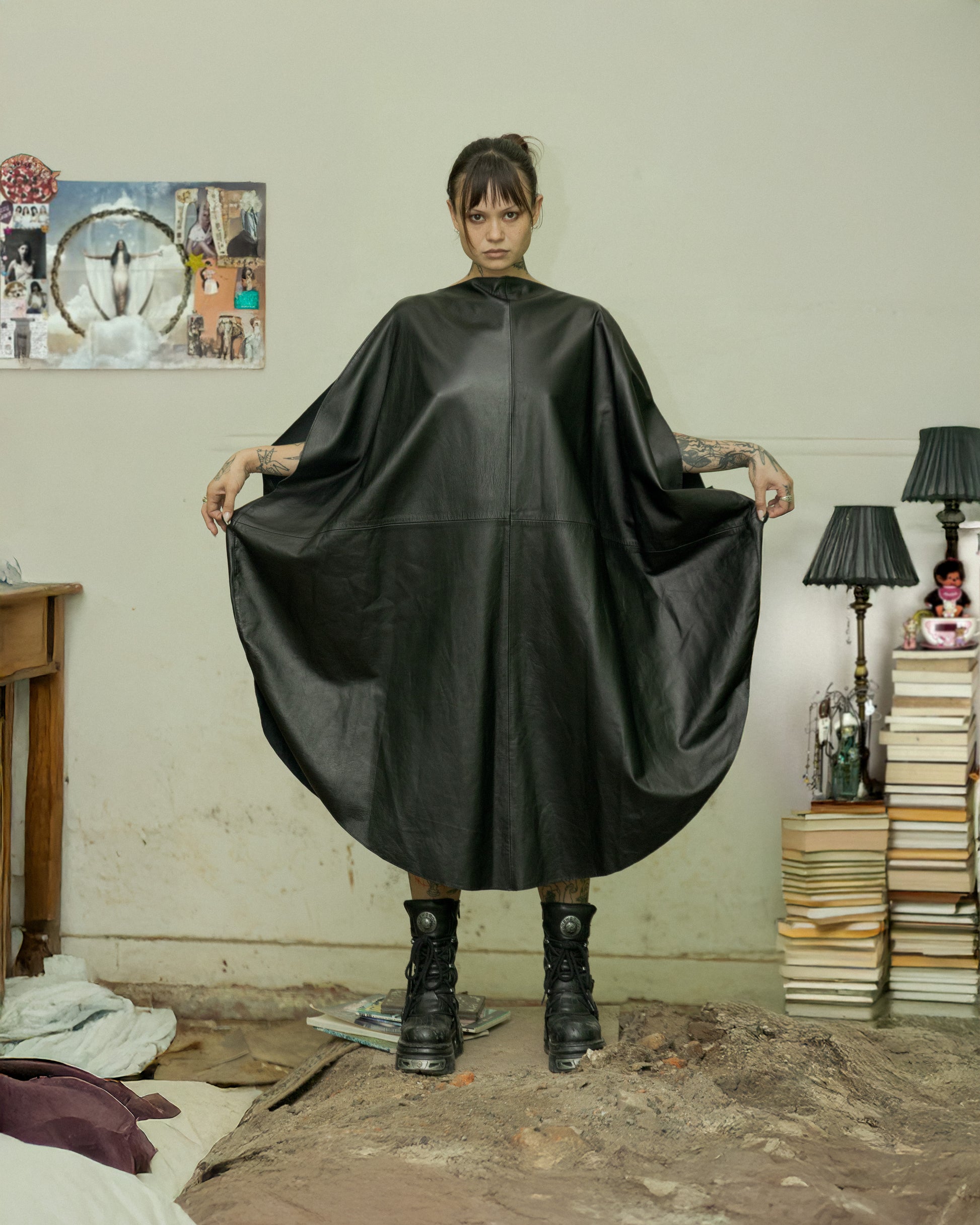 A person standing in a room wearing a black sleeveless, round-shaped lambskin dress with a wide bottom opening, holding the dress away from their body.