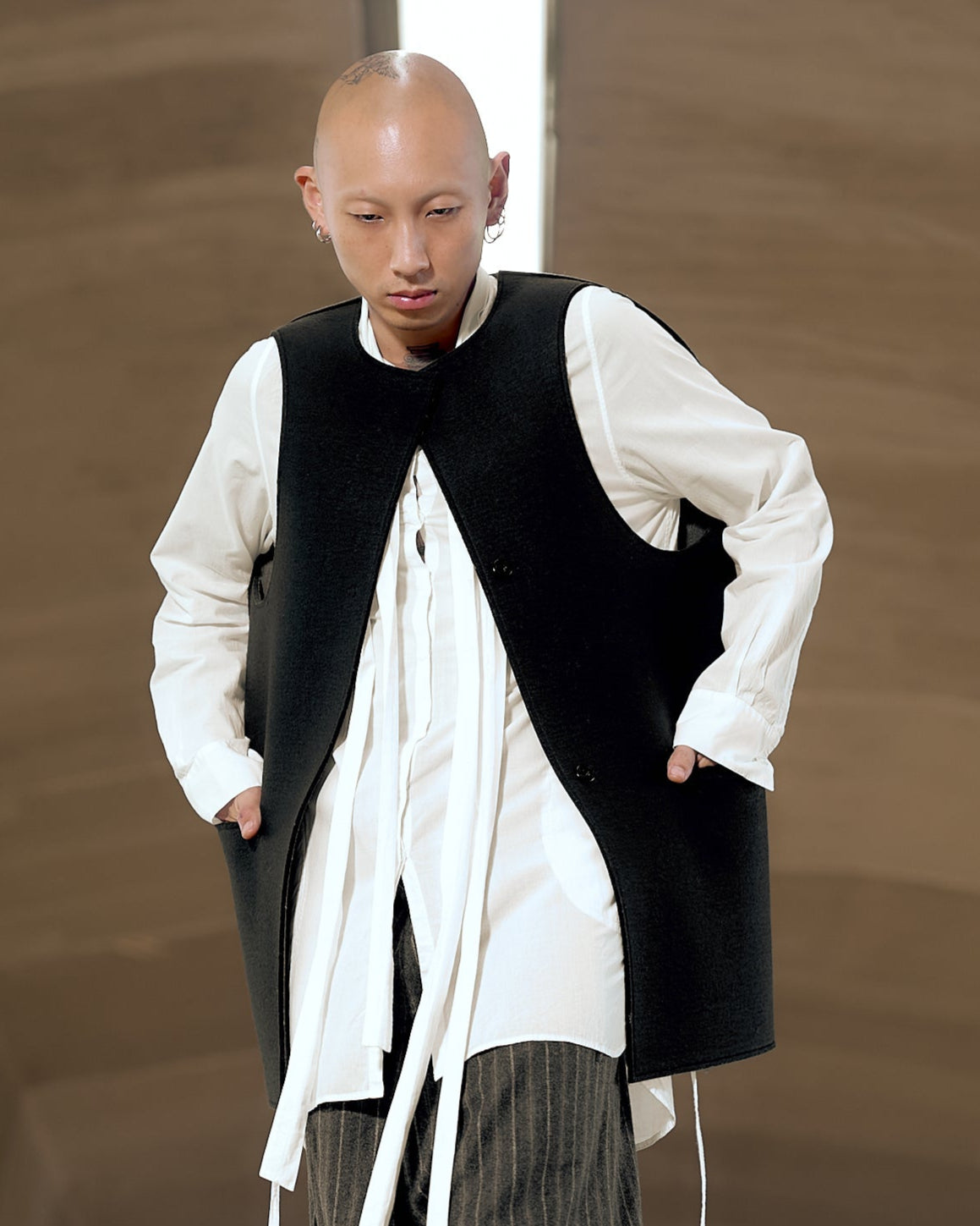 A person in a white shirt and black Maison Margiela vest stands with hands in pockets, wearing pinstriped pants. 