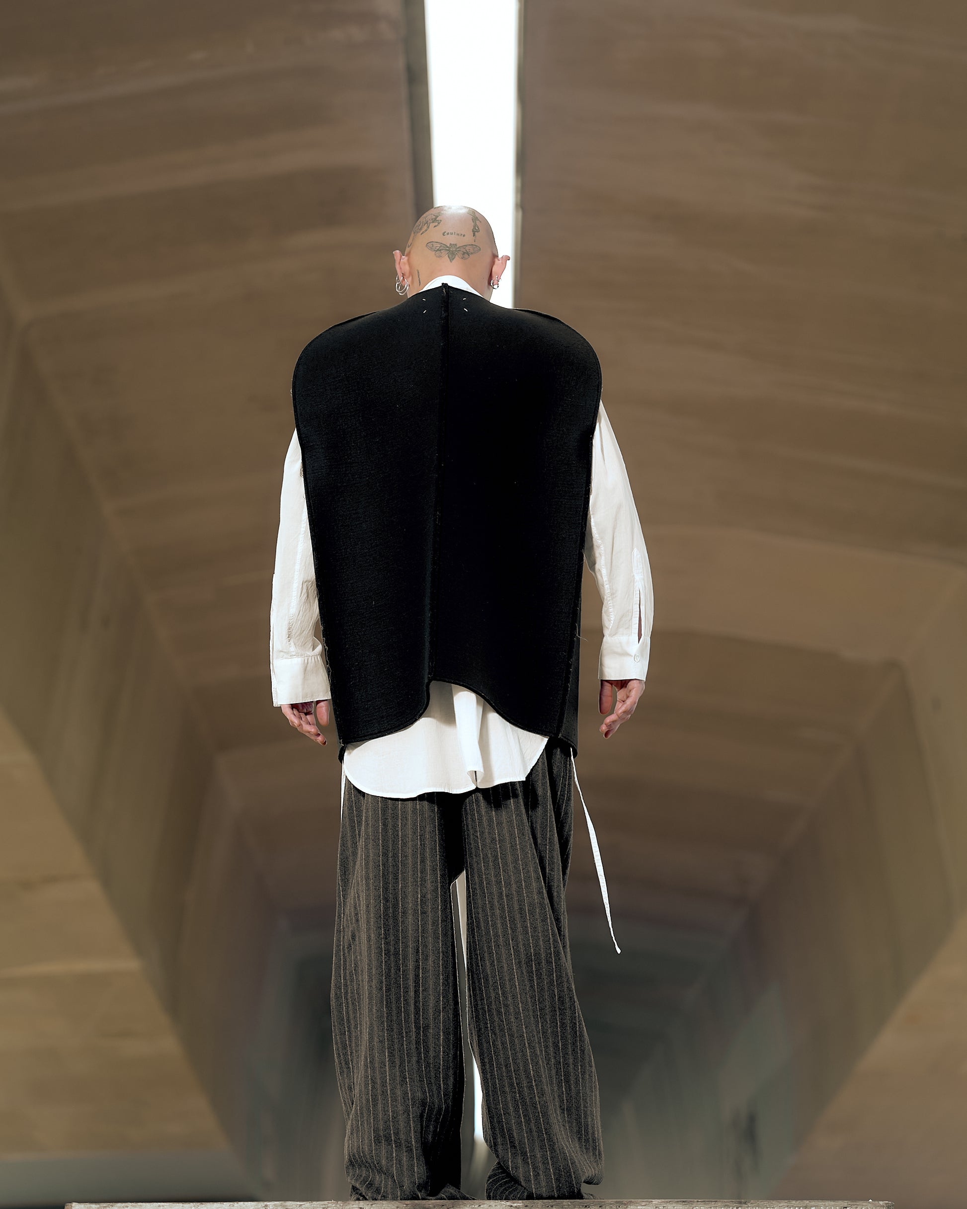 A person stands under an architectural structure, wearing a white shirt, pinstripe pants, and a black Maison Margiela vest. 