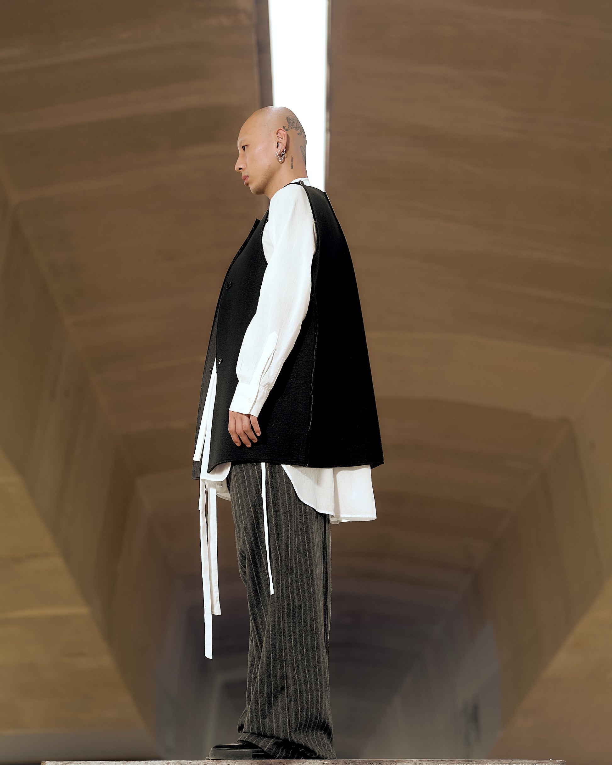 A person stands in profile, wearing a long black Maison Margiela vest over a white shirt with flowing ties and pinstriped pants. The background is an arched tunnel.