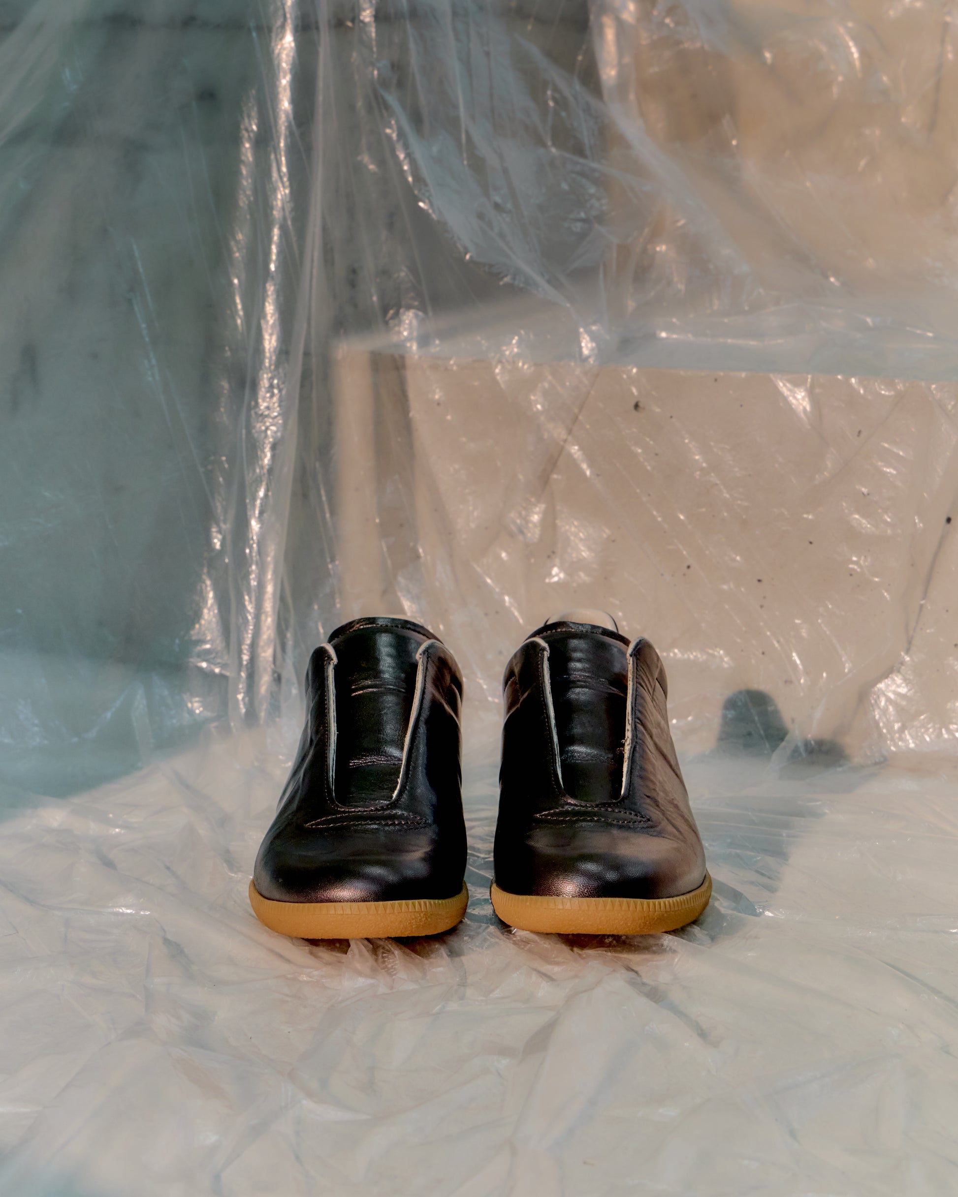 Pair of black Maison Margiela shoes with brown soles on a white surface with plastic sheeting.