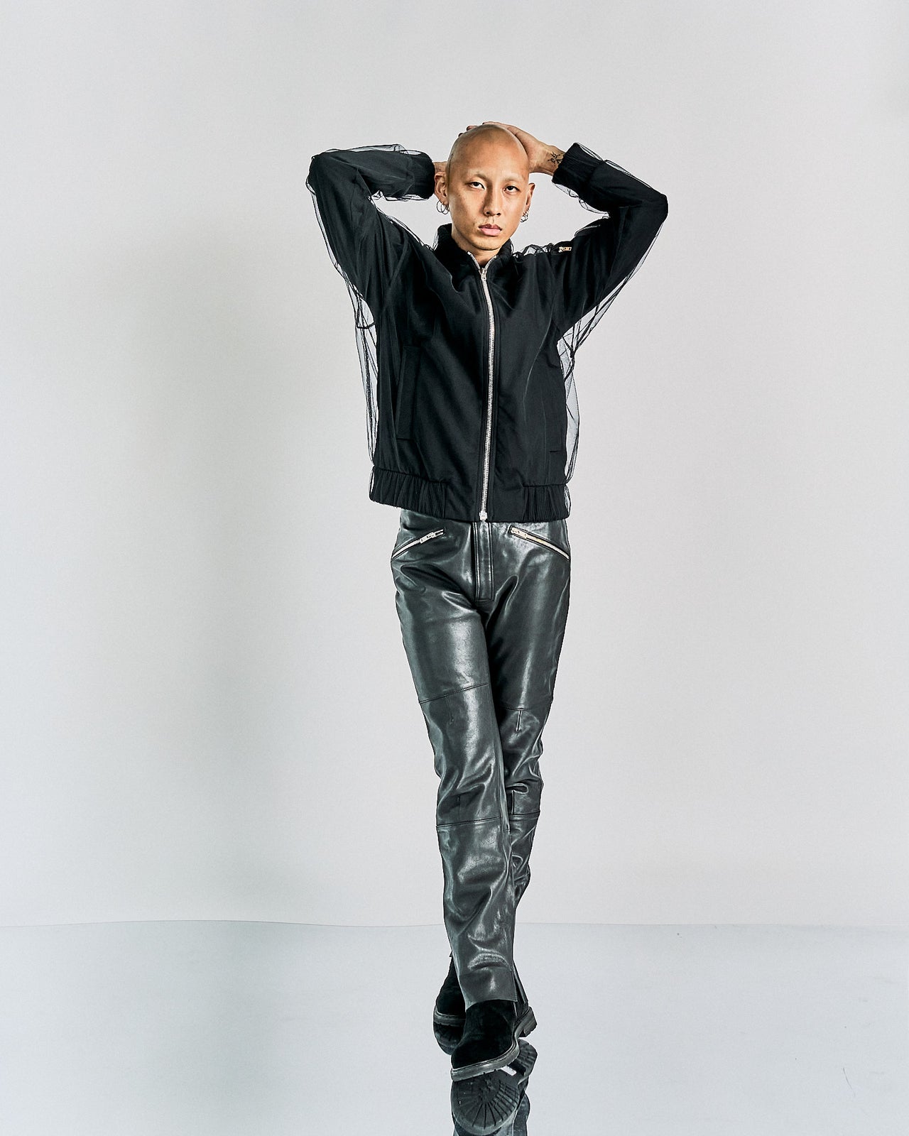 A model wearing a black Maison Margiela bomber jacket with a tulle overlay and ribbed finishings, paired with black leather pants. The model has a shaved head and is posing with one hand to the side.