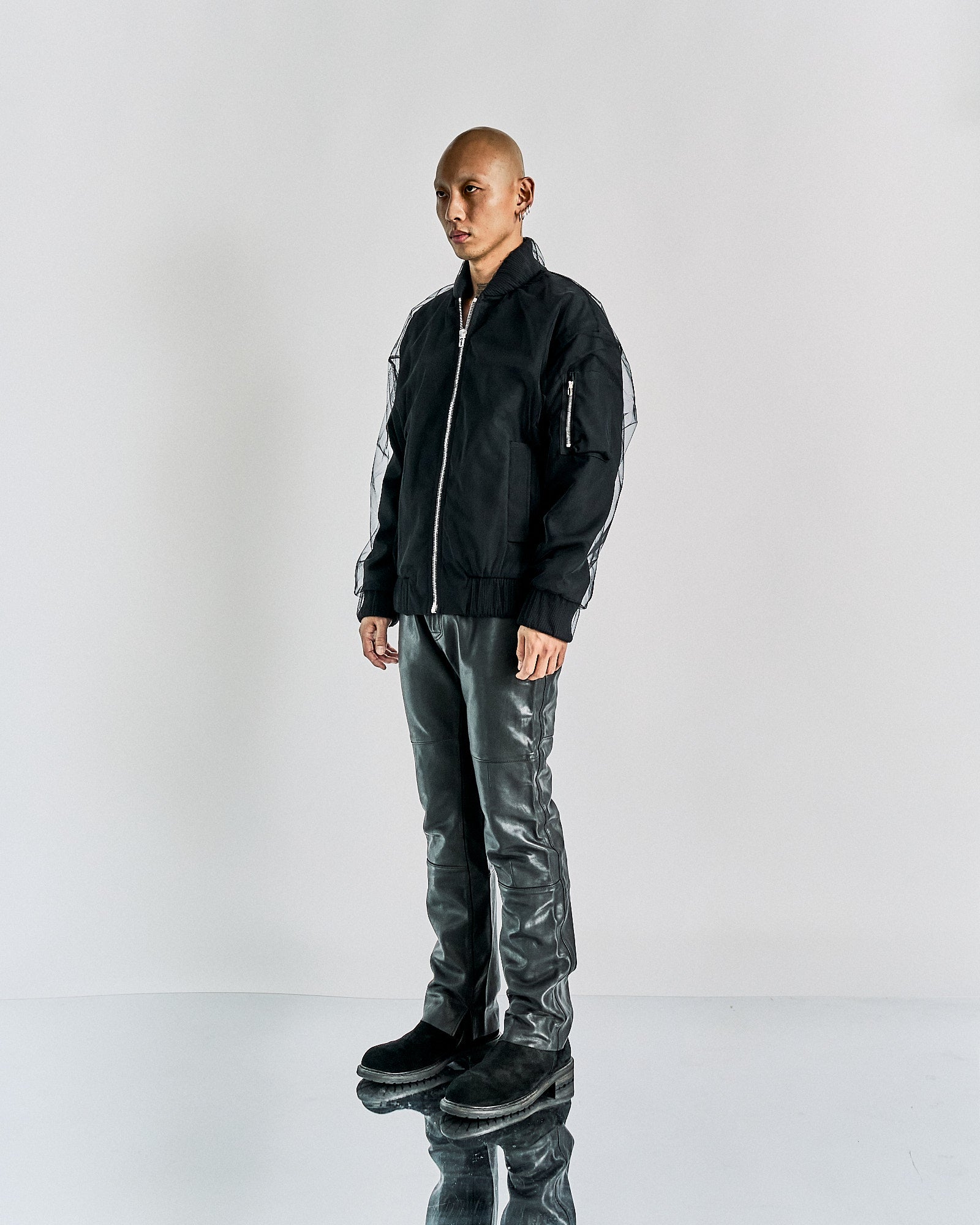 A person stands against a plain background wearing a black zip-up Maison Margiela jacket and shiny grey pants. The atmosphere is minimalist and modern.