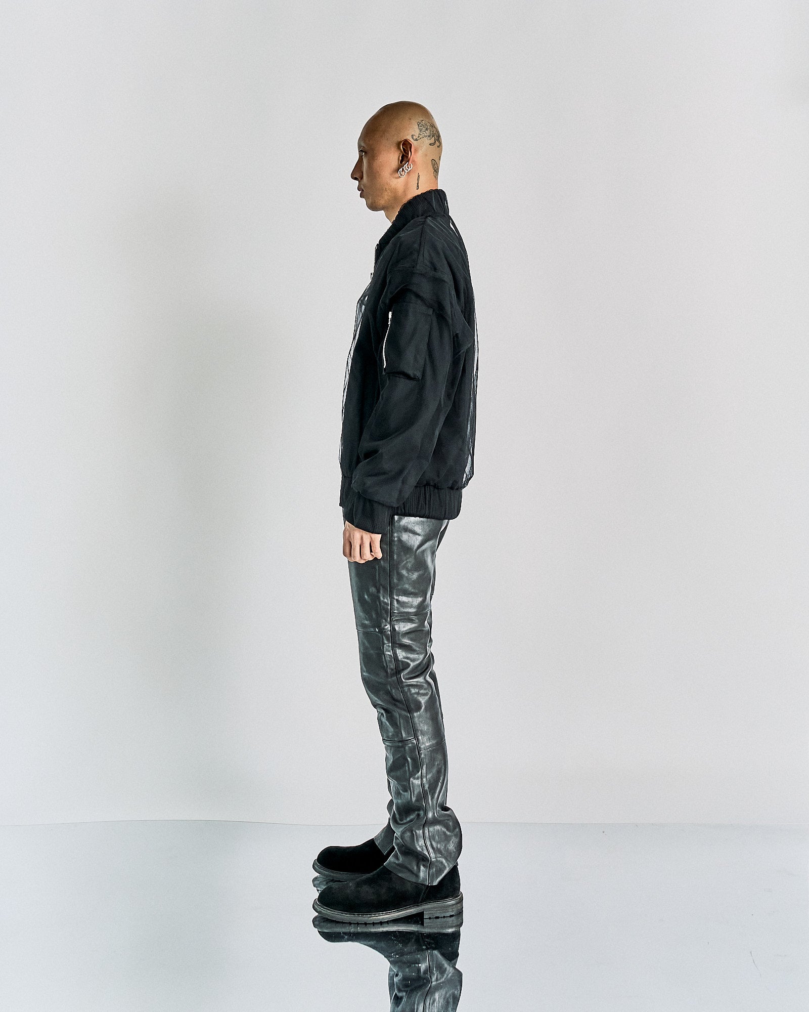 Side profile of a person wearing a black Maison Margiela tulle jacket, leather pants, and boots. The head is shaved and tattooed. 