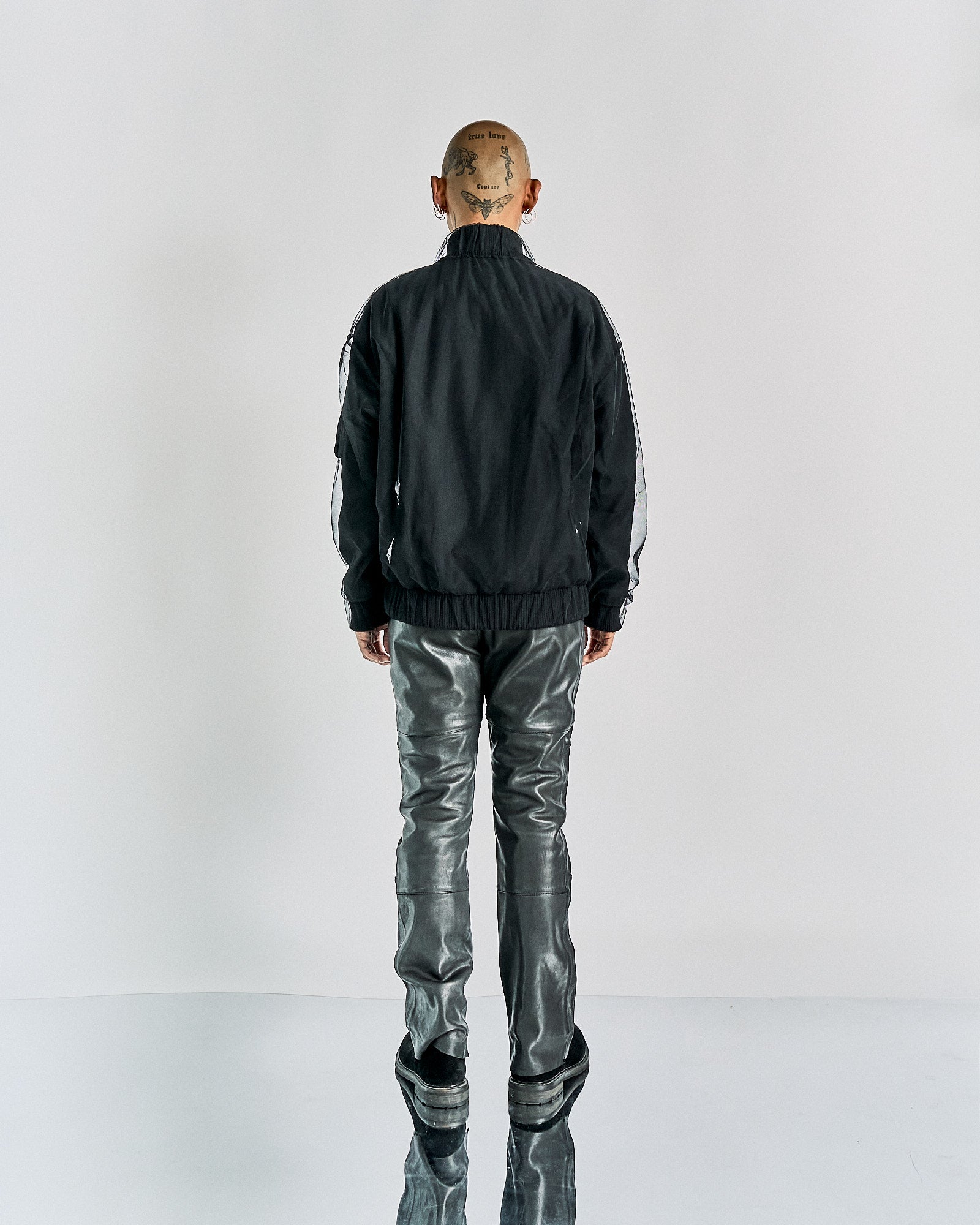 Reverse of a person wearing a black Maison Margiela tulle jacket, leather pants, and boots. The head is shaved and tattooed.