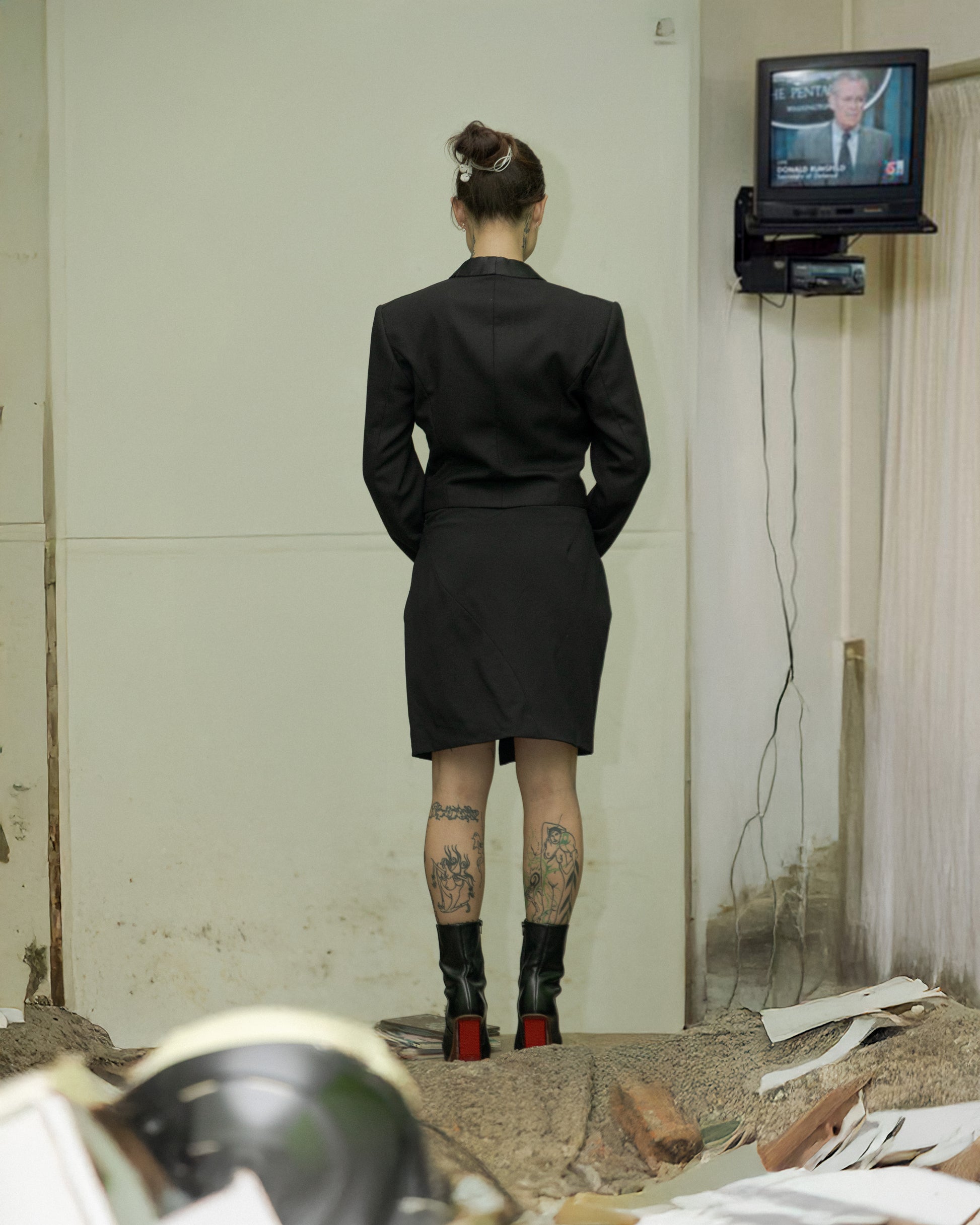 A person viewed from behind wearing a black Maison Margiela tuxedo jacket with a knee-length skirt, standing in a room with various items on the floor and a TV mounted on the wall showing a video.
