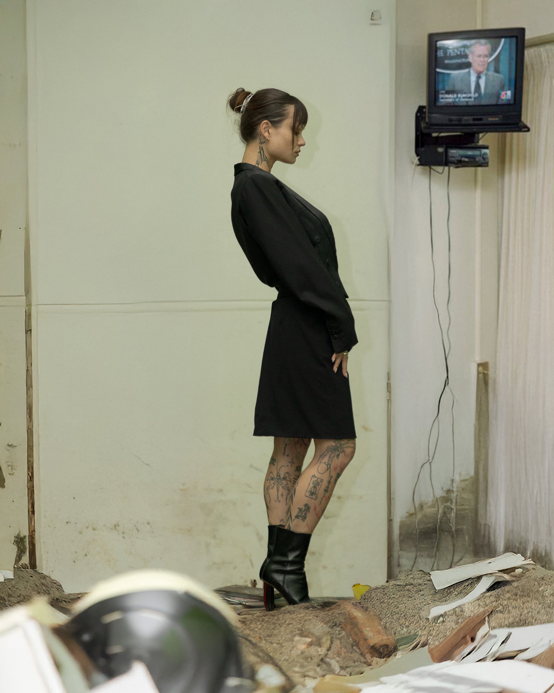 Person in a black Maison Margiela outfit standing in a room with a television mounted on the wall.