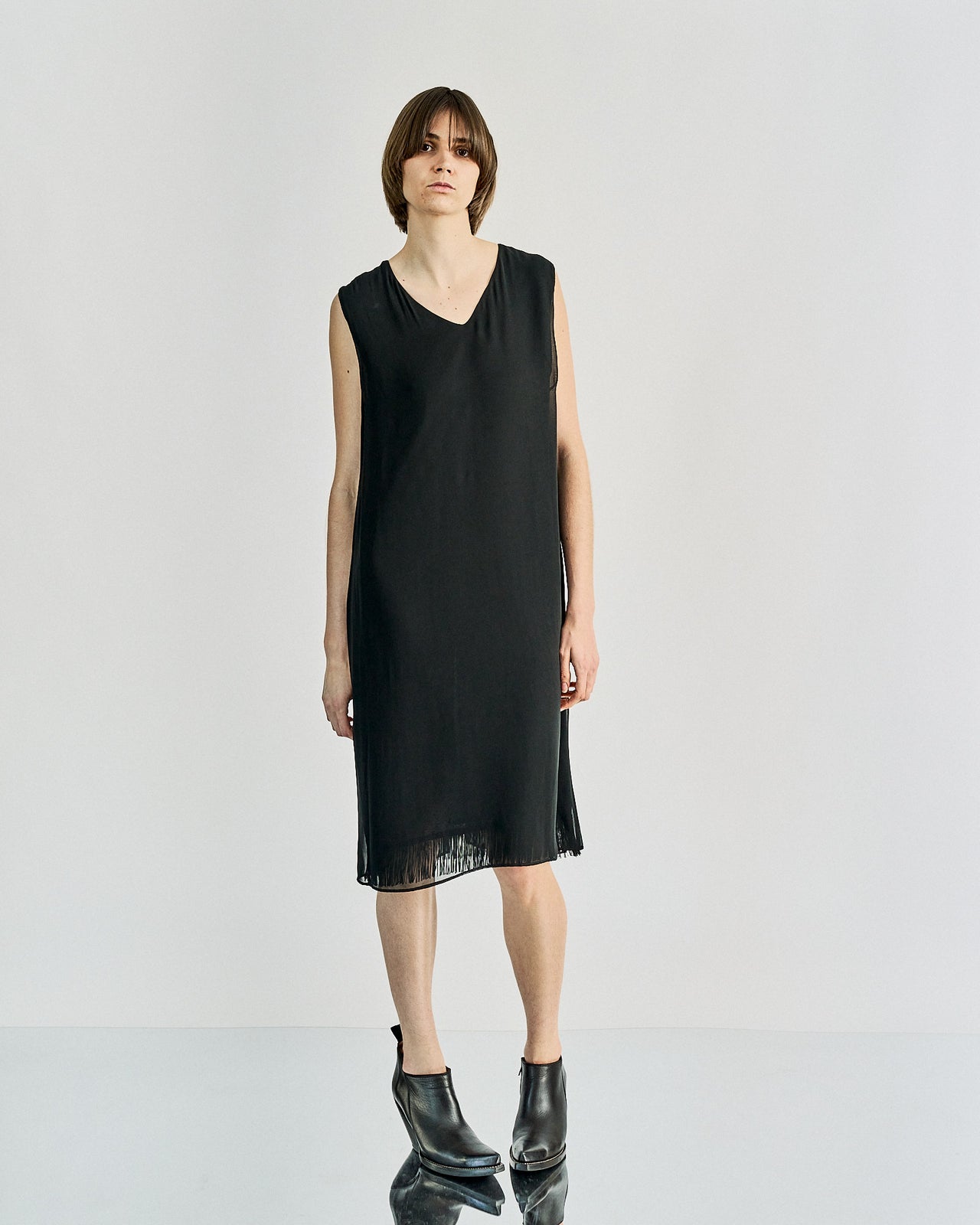 A woman standing wearing a black, V-neck, Maison Margiela tassel-detailed slip dress with a split open back.