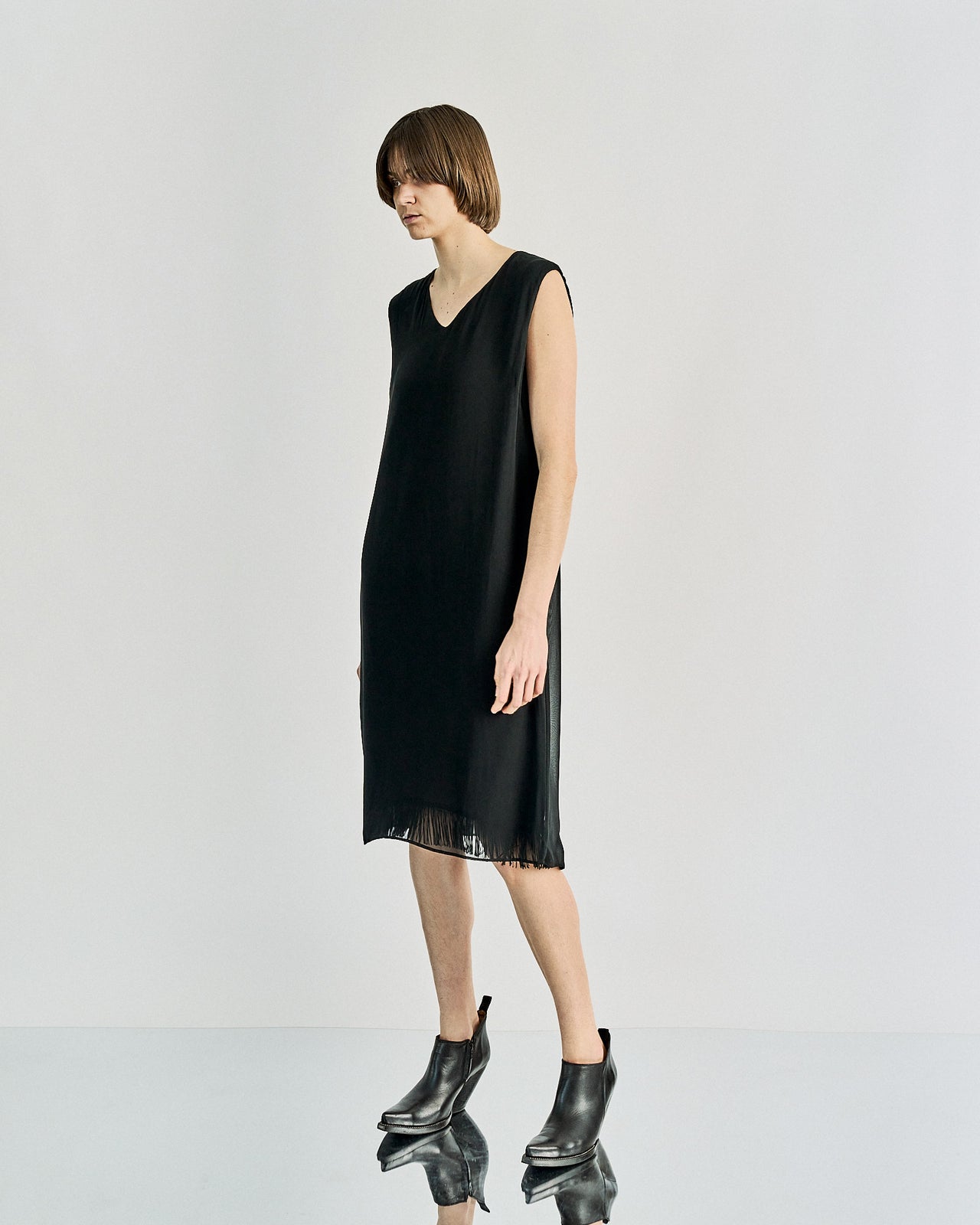 A woman standing wearing a black, V-neck, Maison Margiela tassel-detailed slip dress with a split open back.