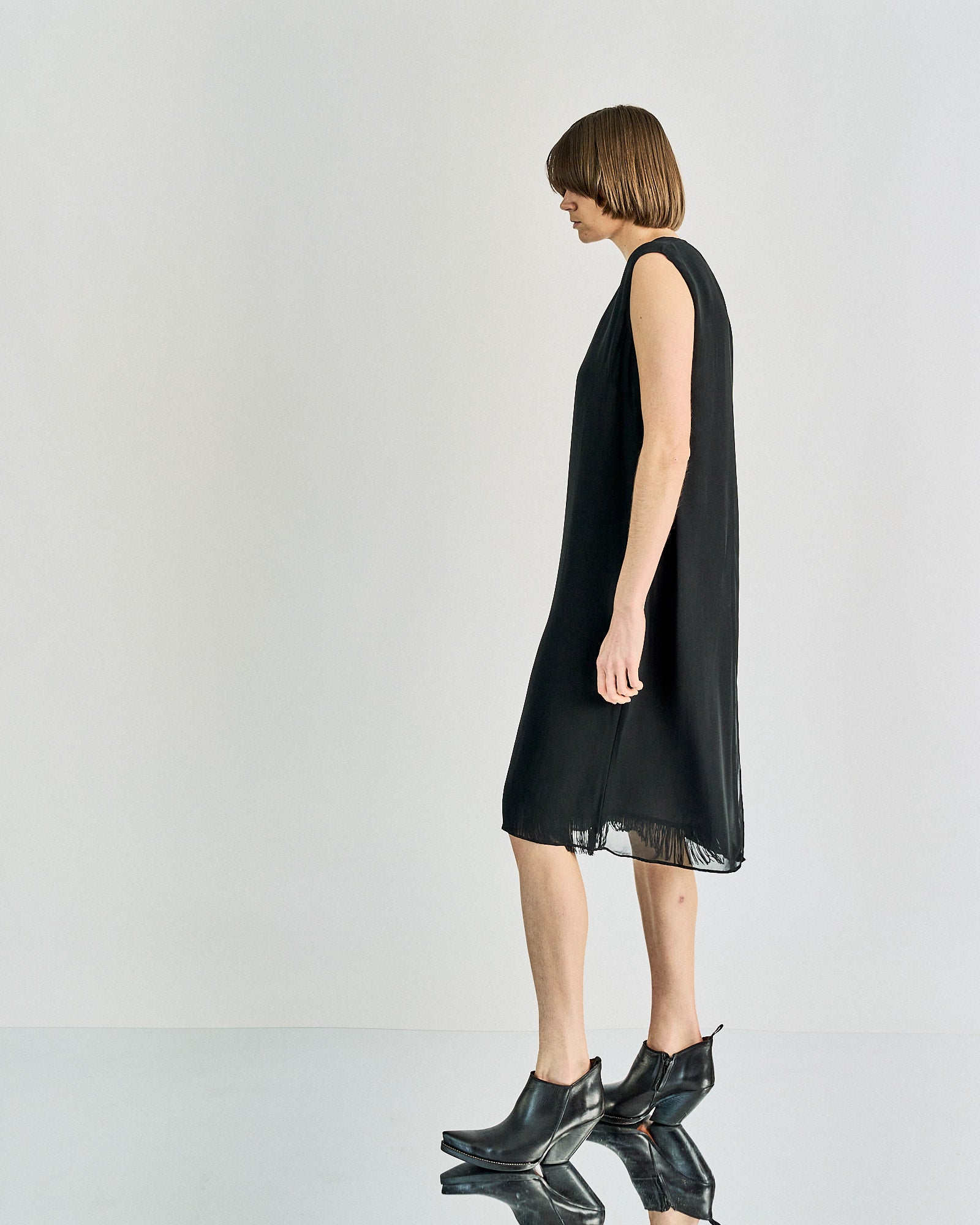 A person in a sleeveless black Maison Margiela dress and short brown hair stands against a plain background, wearing black ankle boots