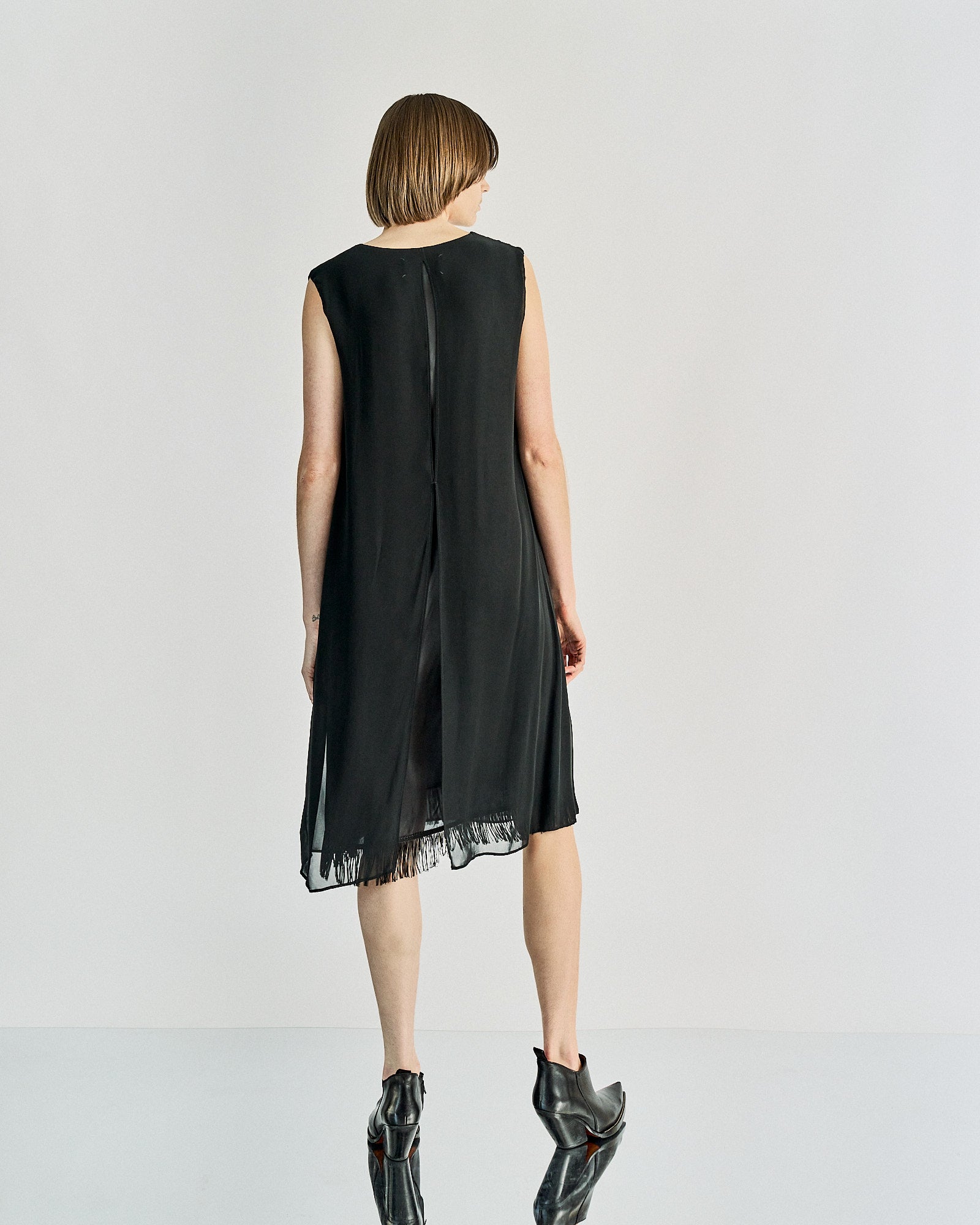 A person stands facing away, wearing a sleeveless black Maison Margiela dress with fringe at the hem. The setting is minimal, with a neutral background.