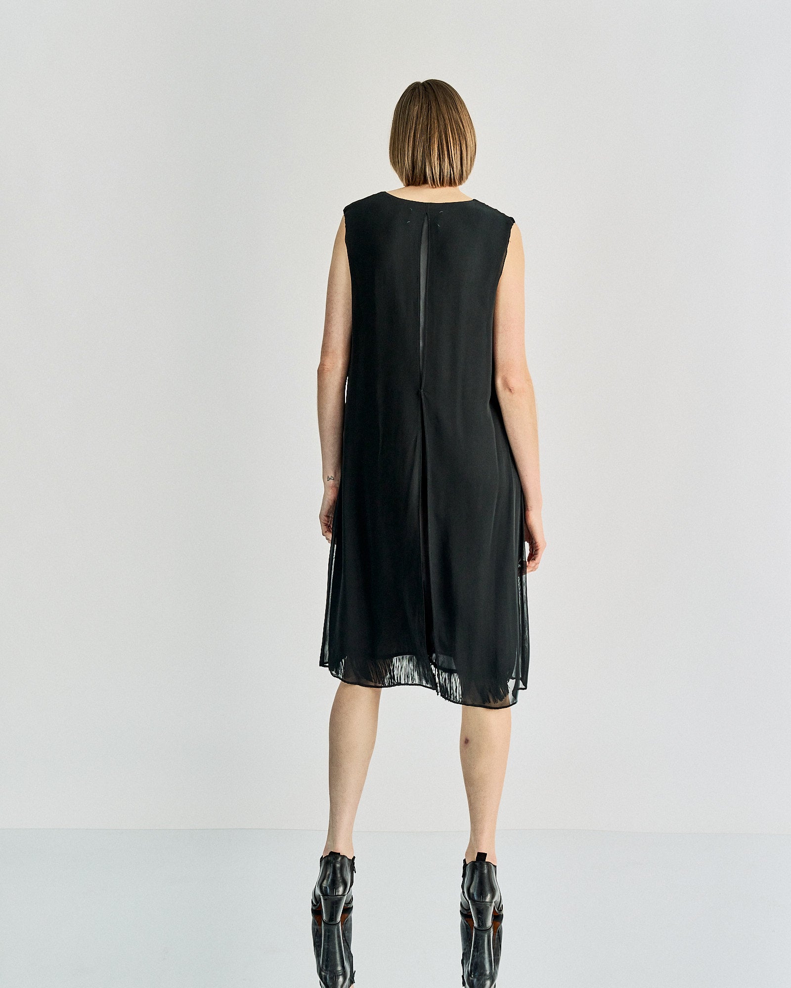 A person stands facing away, wearing a sleeveless black Maison Margiela dress with fringe at the hem. The setting is minimal, with a neutral background.