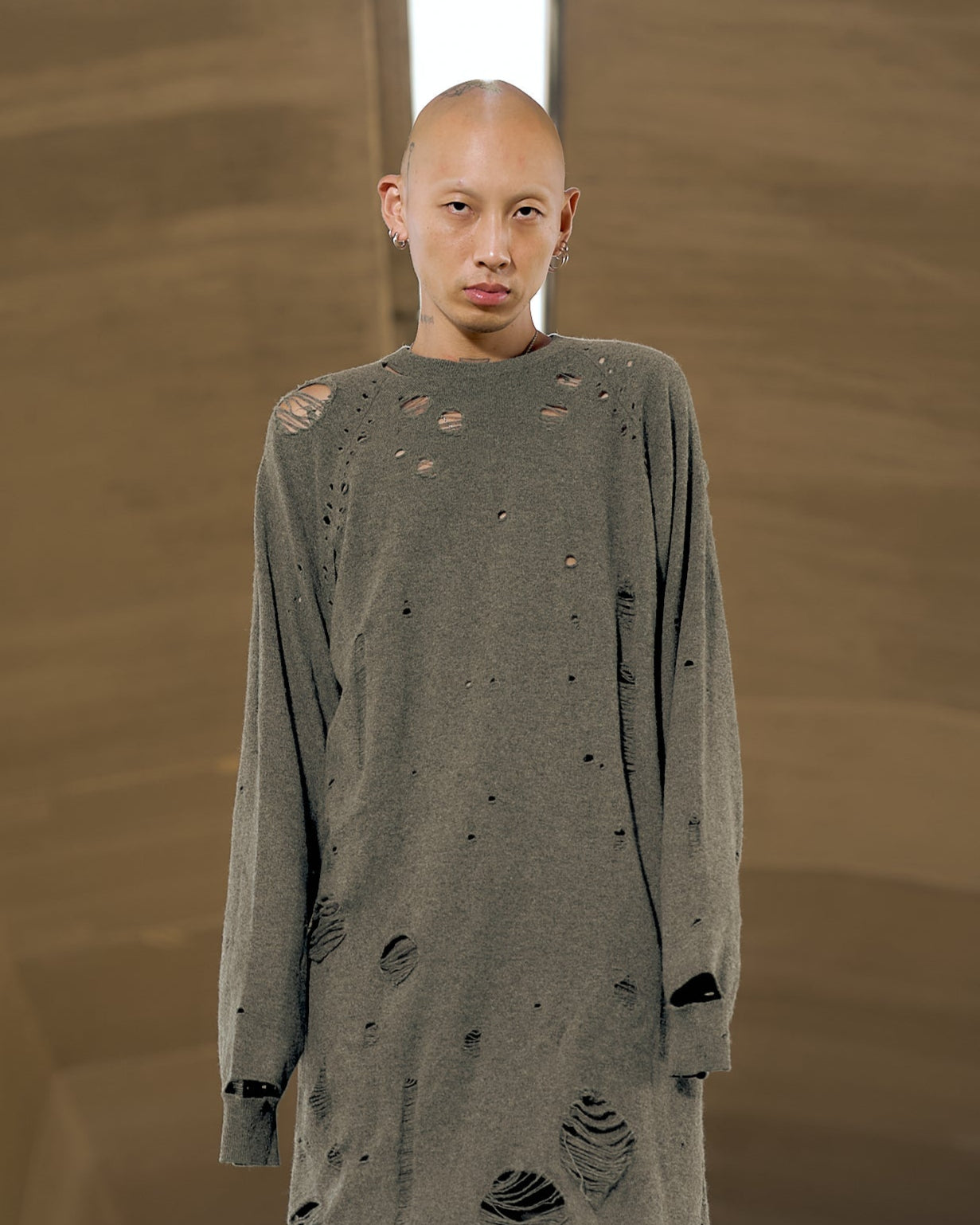 A person stands confidently, wearing an oversized gray Maison Margiela sweater with many holes, against a neutral brown background.