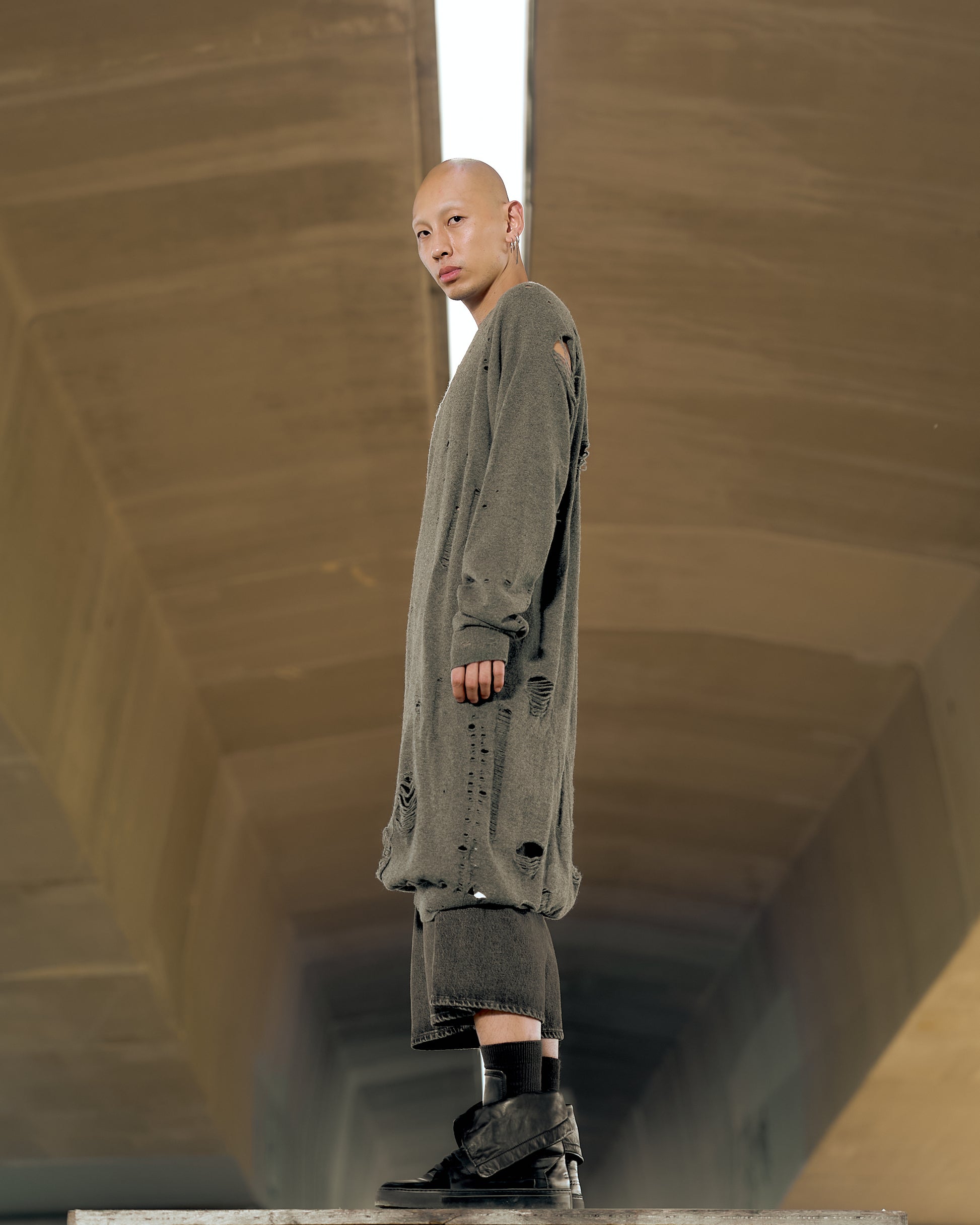 A person stands confidently, wearing an oversized gray Maison Margiela sweater with many holes, against a neutral brown background. Their expression is calm and composed.