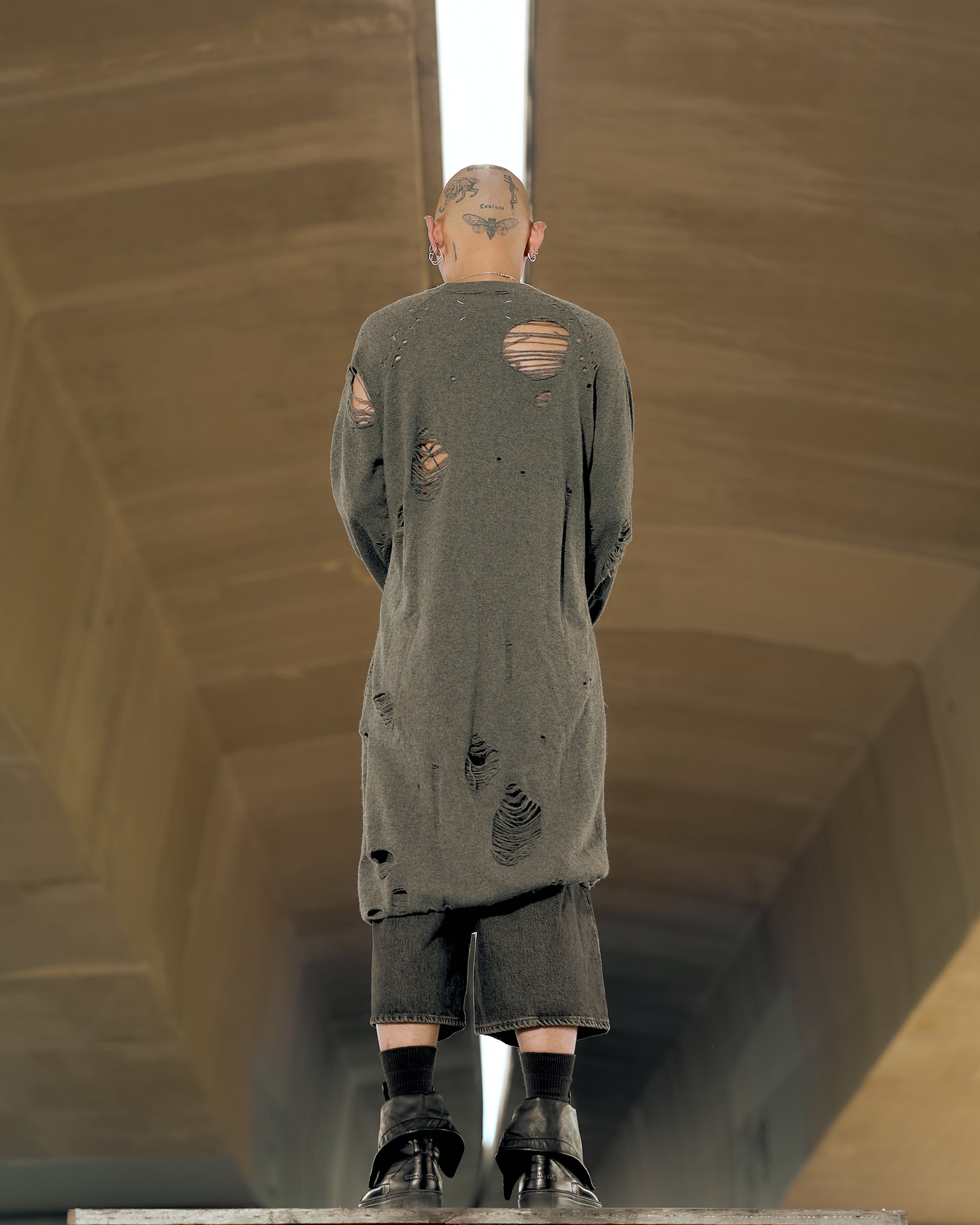 Bald person with head tattoos, facing away, wearing a distressed gray Maison Margiela sweater and black shorts in an urban setting, conveying a grunge aesthetic.