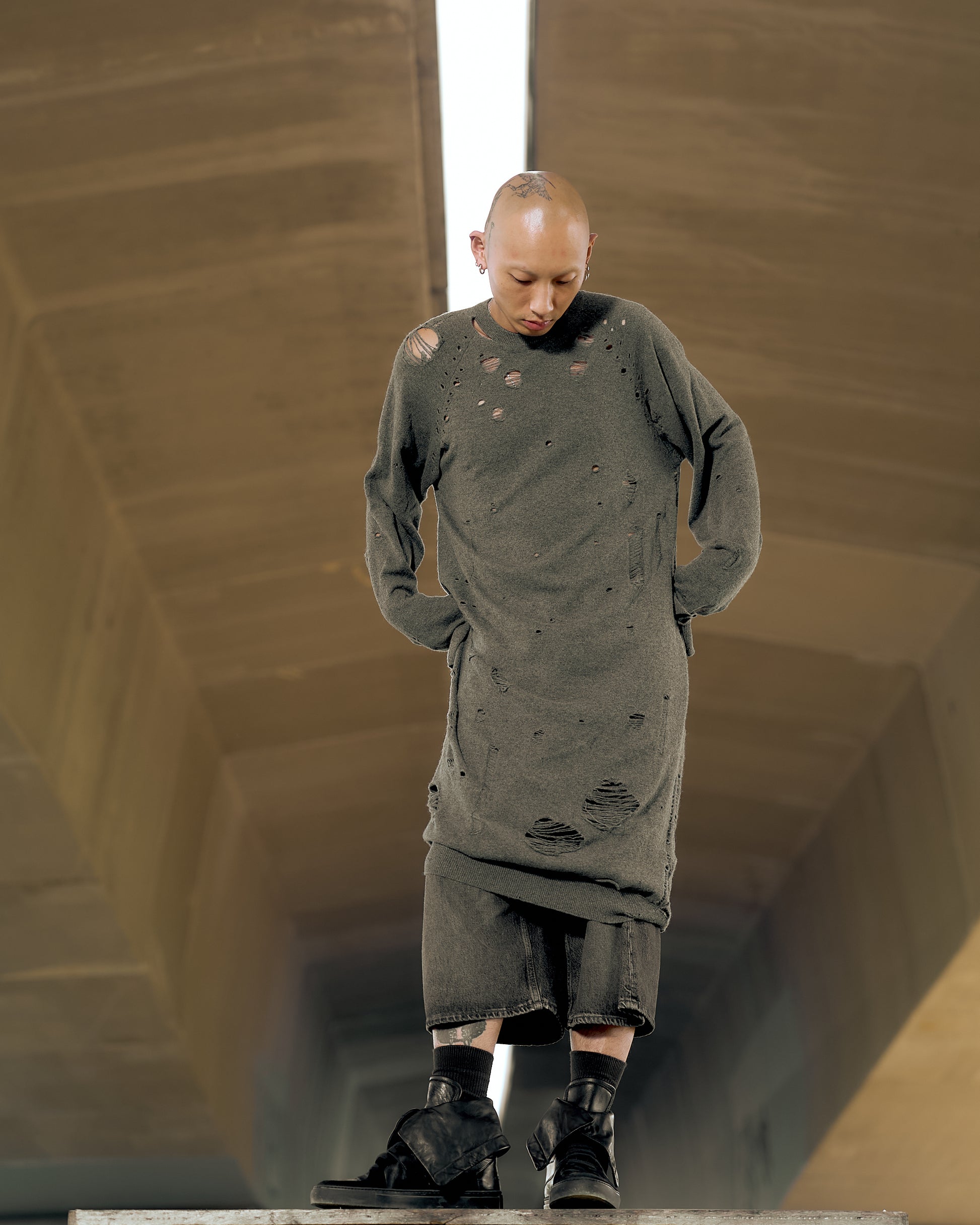 A person stands with eyes downcast, wearing a long, distressed gray Maison Margiela sweater and dark boots. 