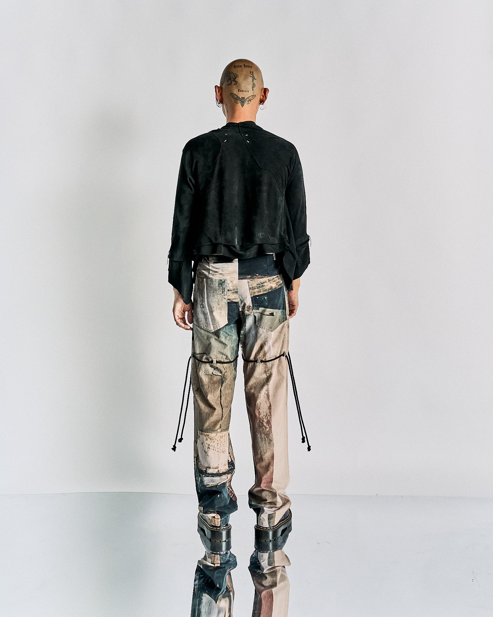 Man standing with back to camera, wearing a black Maison Margiela jacket and patchwork pants.