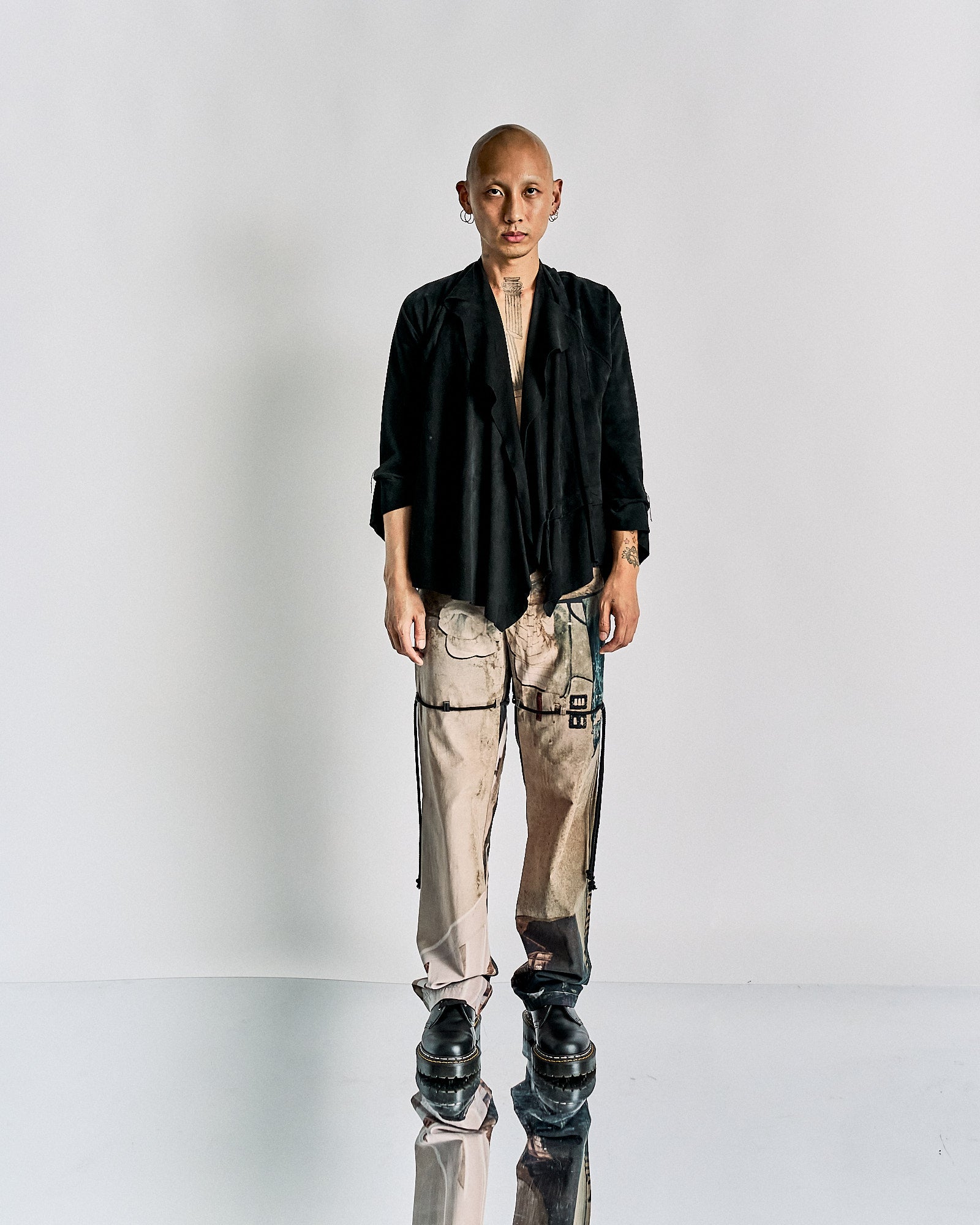 A person stands in a minimalistic setting, wearing a black Maison Margiela jacket, beige cargo pants, and black boots.
