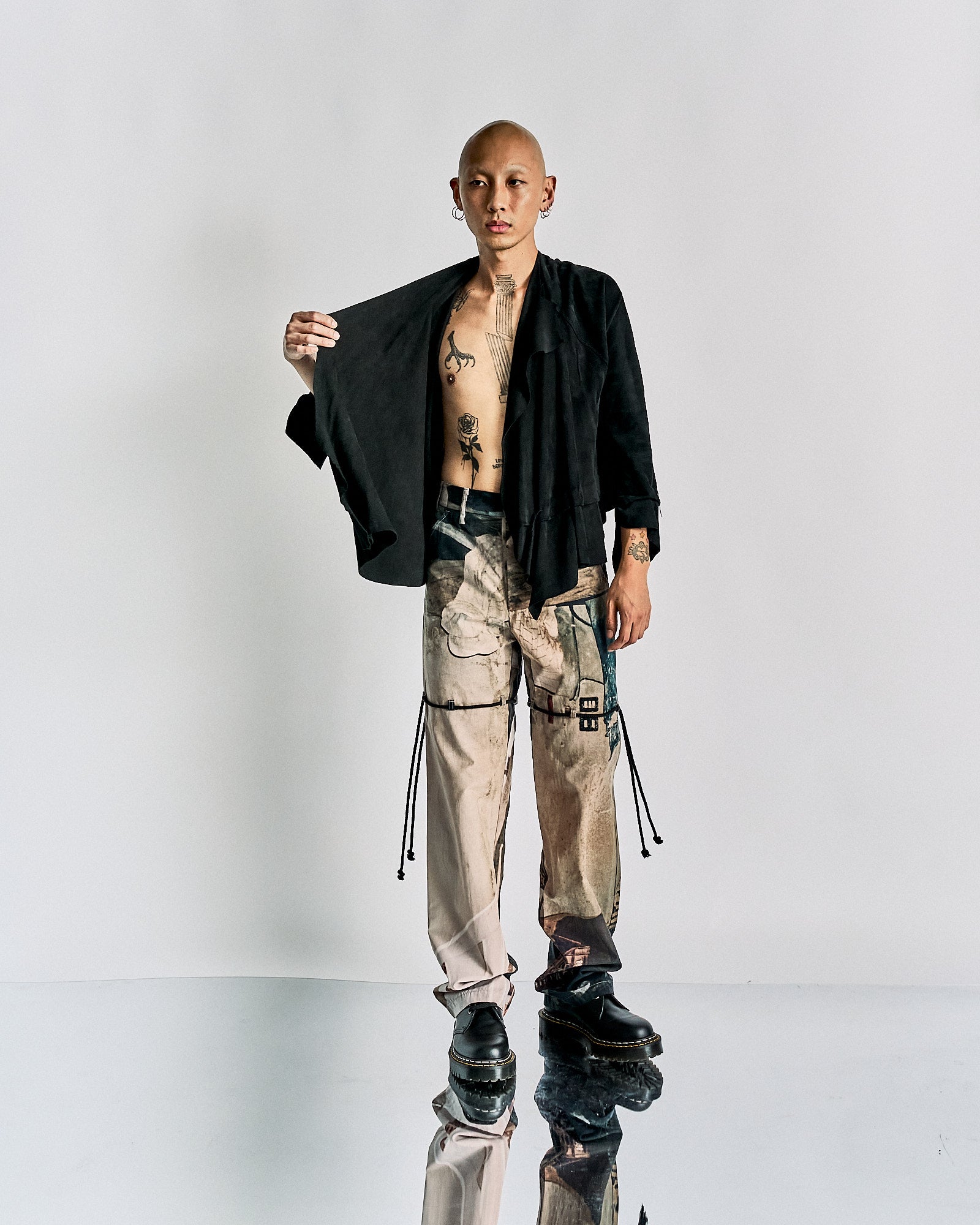 A person stands against a white background, wearing patterned trousers, a black open Maison Margiela jacket, and polished black shoes.