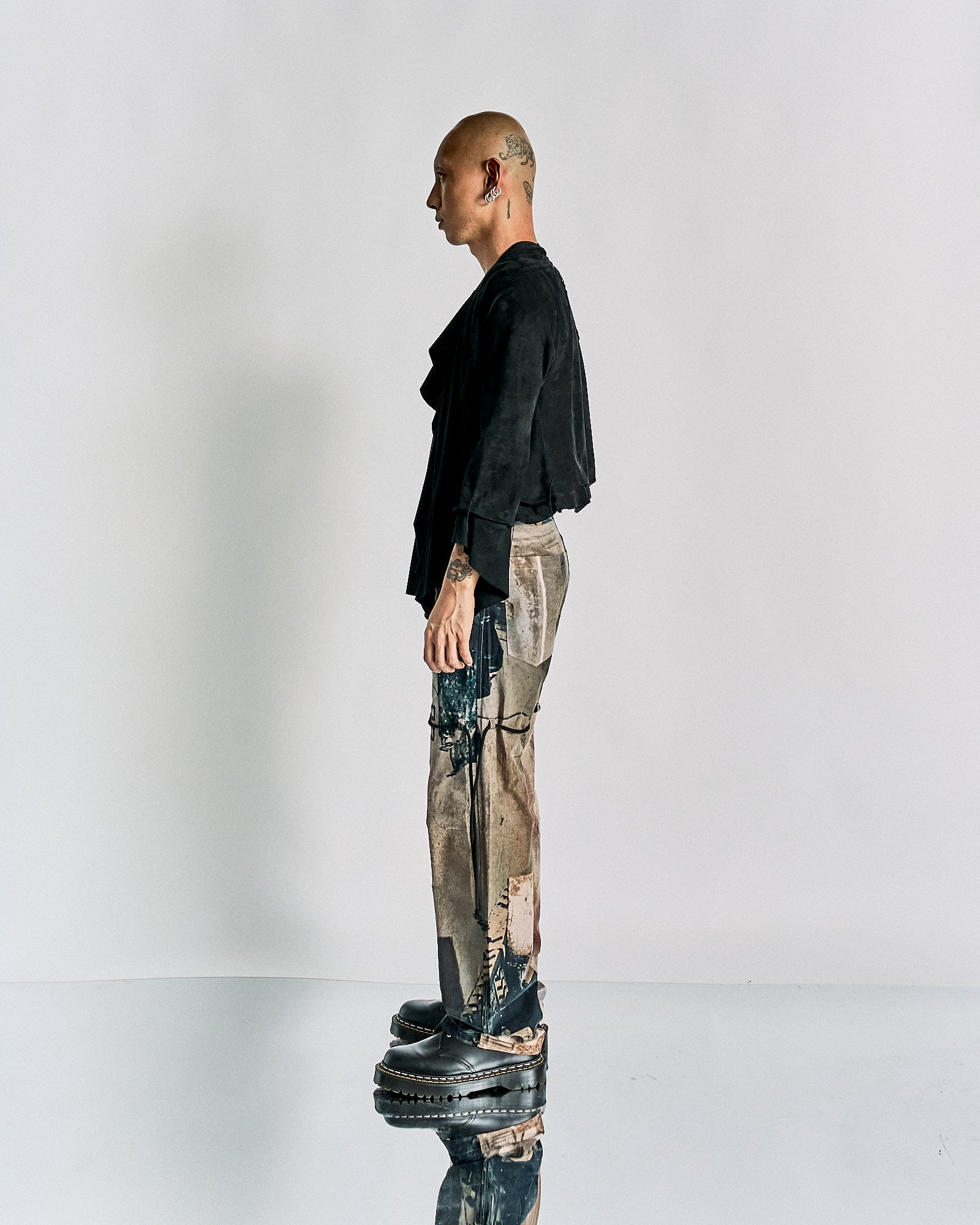 A person stands in profile against a white background, wearing a black Maison Margiela jacket, worn-out patterned pants, and platform shoes. 
