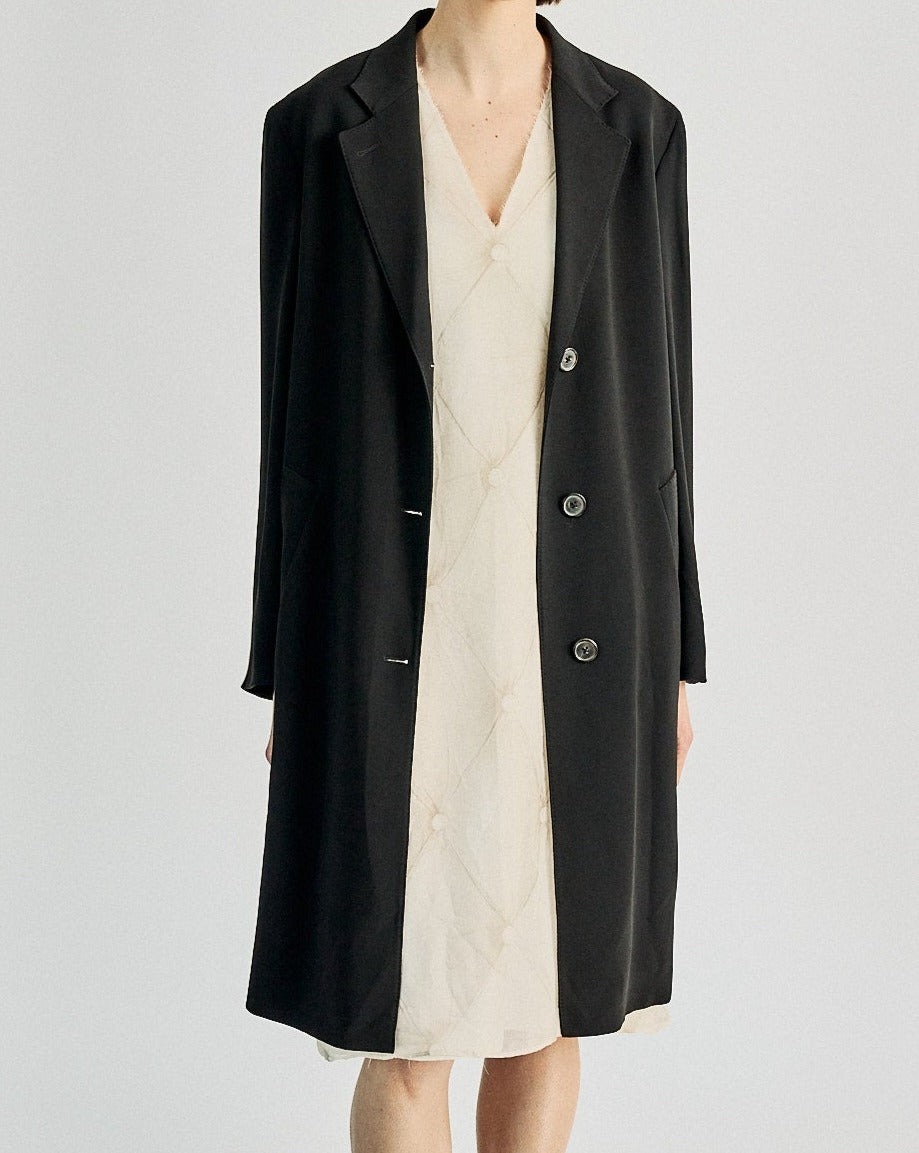 Person wearing a long black Maison Margiela coat over a cream quilted dress. The style is minimalist, with a refined and elegant tone against a plain background.