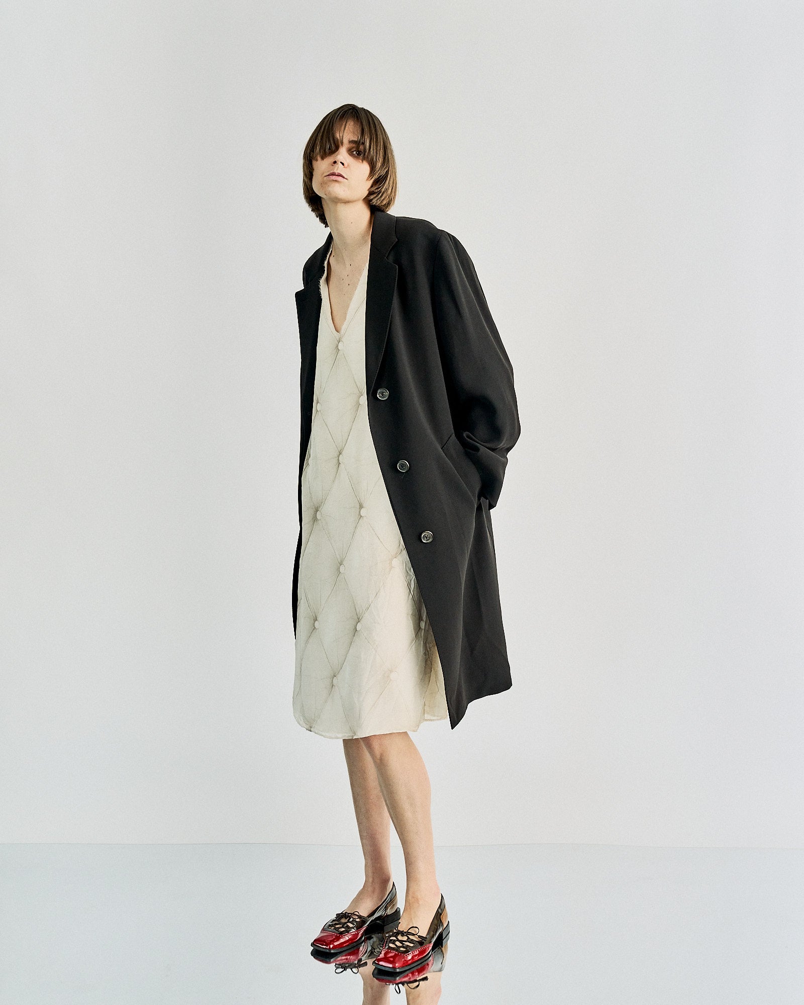 Fashionable individual in a black Maison Margiela coat over a cream quilted dress, red and black patent shoes. The look is modern and chic against a minimalist backdrop.