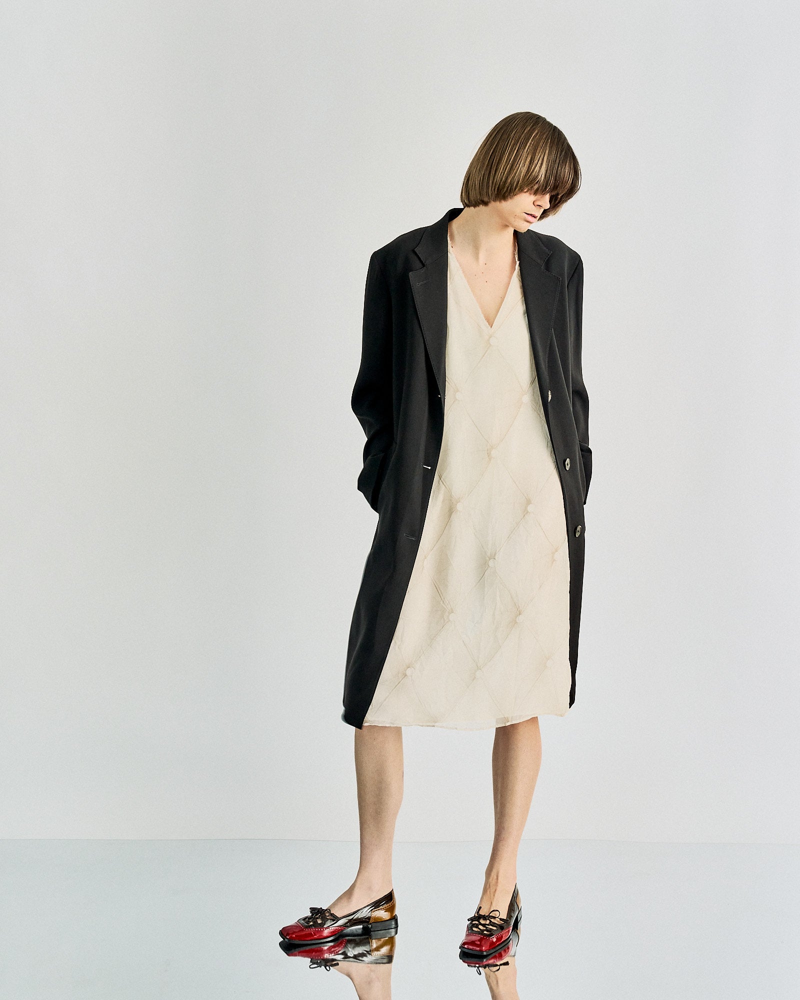Fashionable individual in a black Maison Margiela coat over a cream quilted dress, red and black patent shoes. The look is modern and chic against a minimalist backdrop.