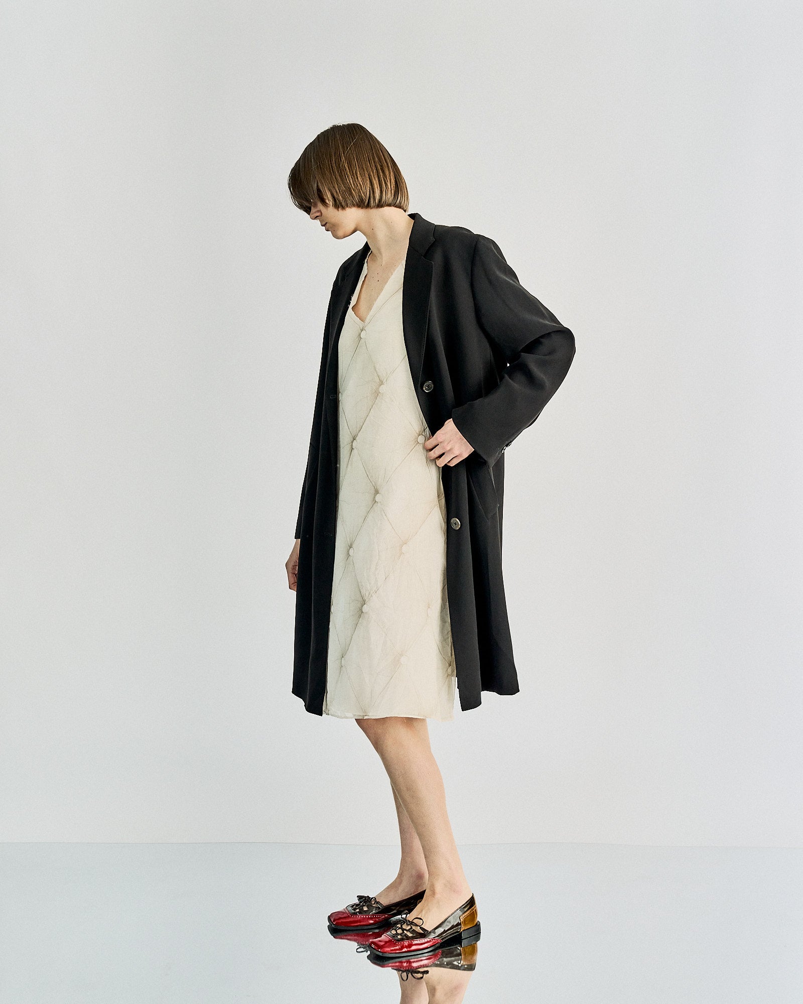 A person with short hair wearing a long black Maison Margiela coat over a white dress, standing against a plain backdrop. They wear red and black shoes, exuding a minimalist and stylish tone.