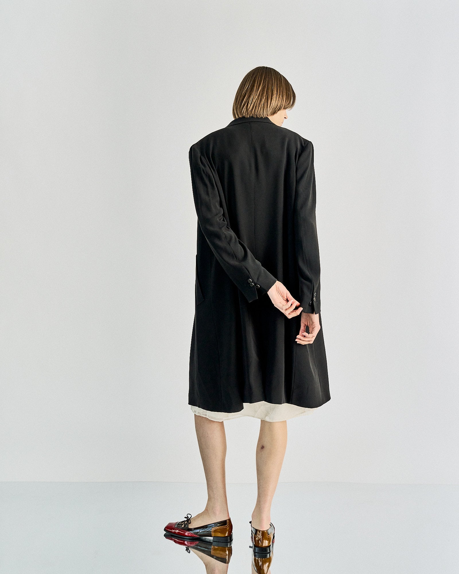 A person stands with their back to the camera, wearing a long Maison Margiela black coat over a white dress. They are pulling at the coat's sleeve. The shoes are colorful with a polished finish