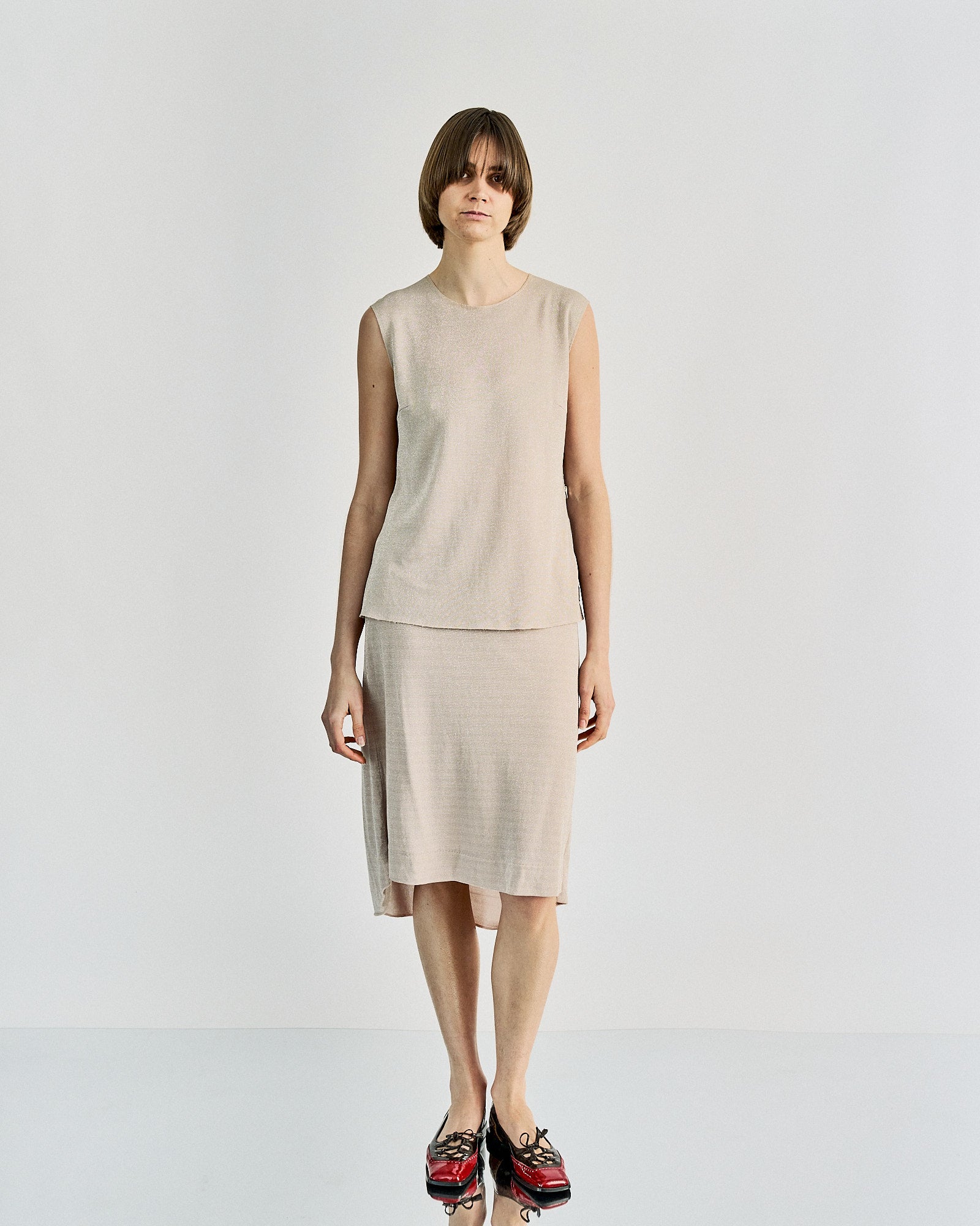 A person with short hair is wearing a sleeveless beige top and matching knee-length skirt from Maison Margiela , standing against a plain white background, looking peaceful.