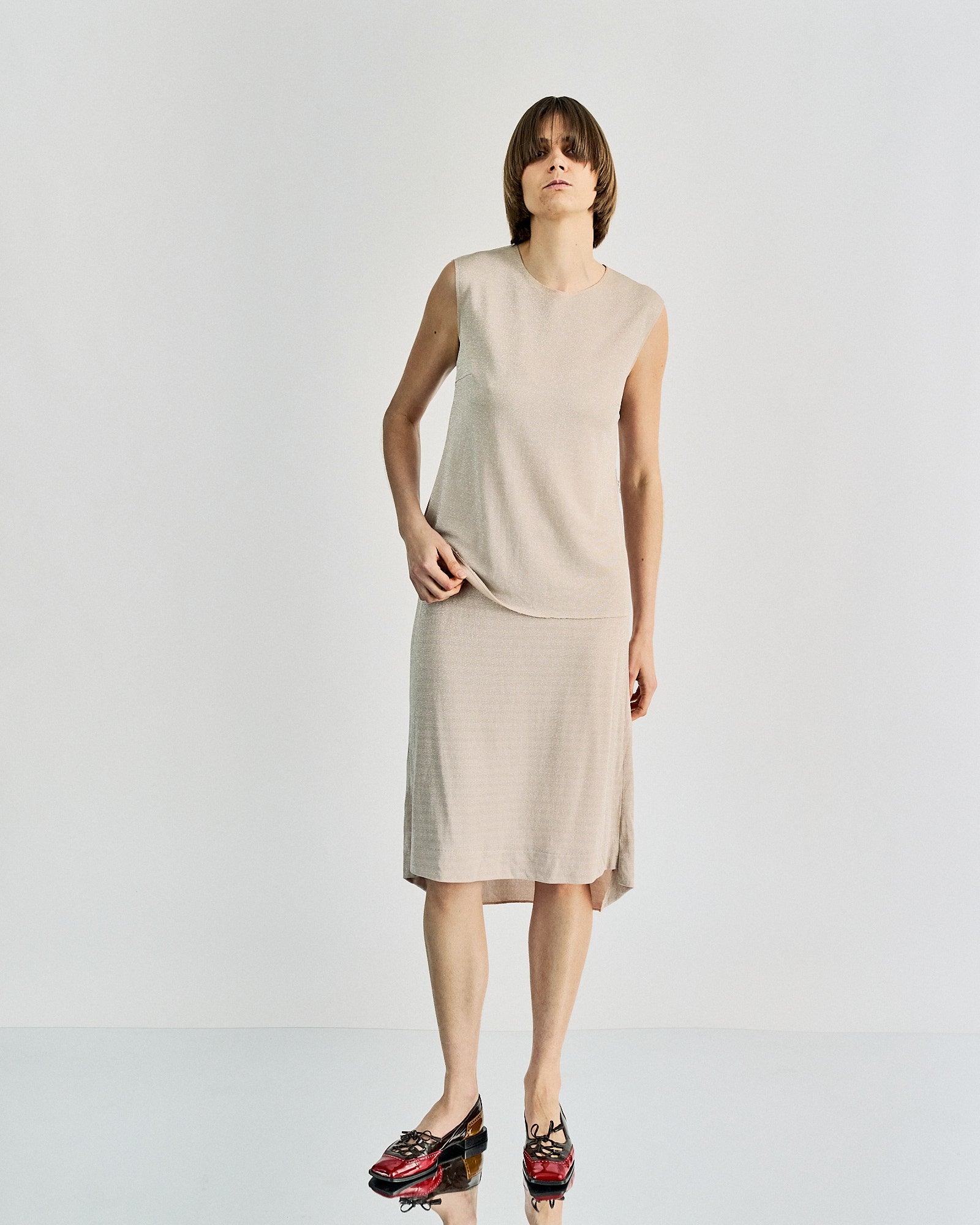 A person wearing a light beige sleeveless Maison Margiela twin set and colourful shoes stands against a plain white background, conveying a relaxed, minimalist style.