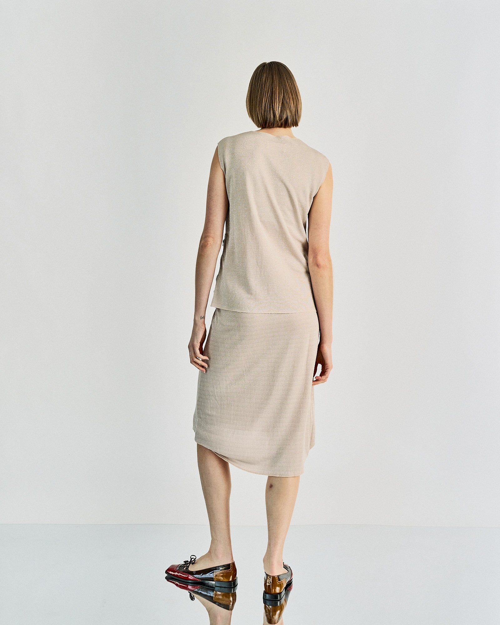 A person stands facing away, wearing a sleeveless beige Maison Margiela top and matching pencil skirt. They have short hair and are wearing shiny multicoloured shoes.