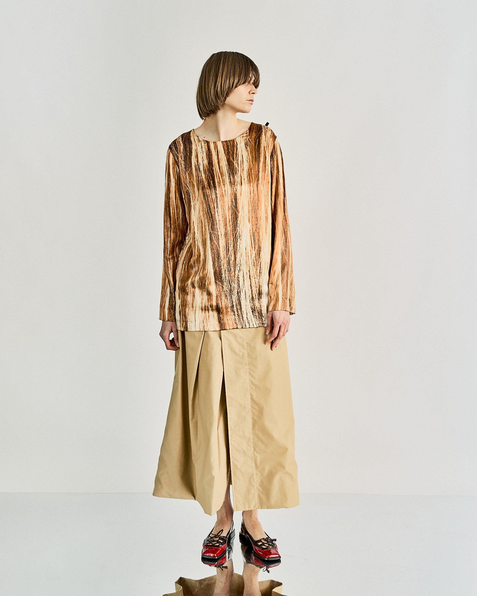 Person in a brown textured Maison Margiela top and beige wide-leg pants stands against a plain background. They wear red and black shoes, exuding a modern, minimalist vibe.