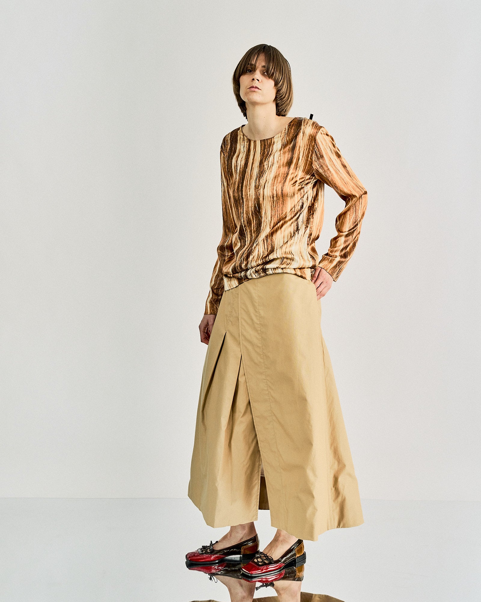 A model in a textured brown long-sleeve Maison Margiela top and a beige maxi skirt with a front slit, wearing glossy black and red shoes, stands confidently on a reflective floor.