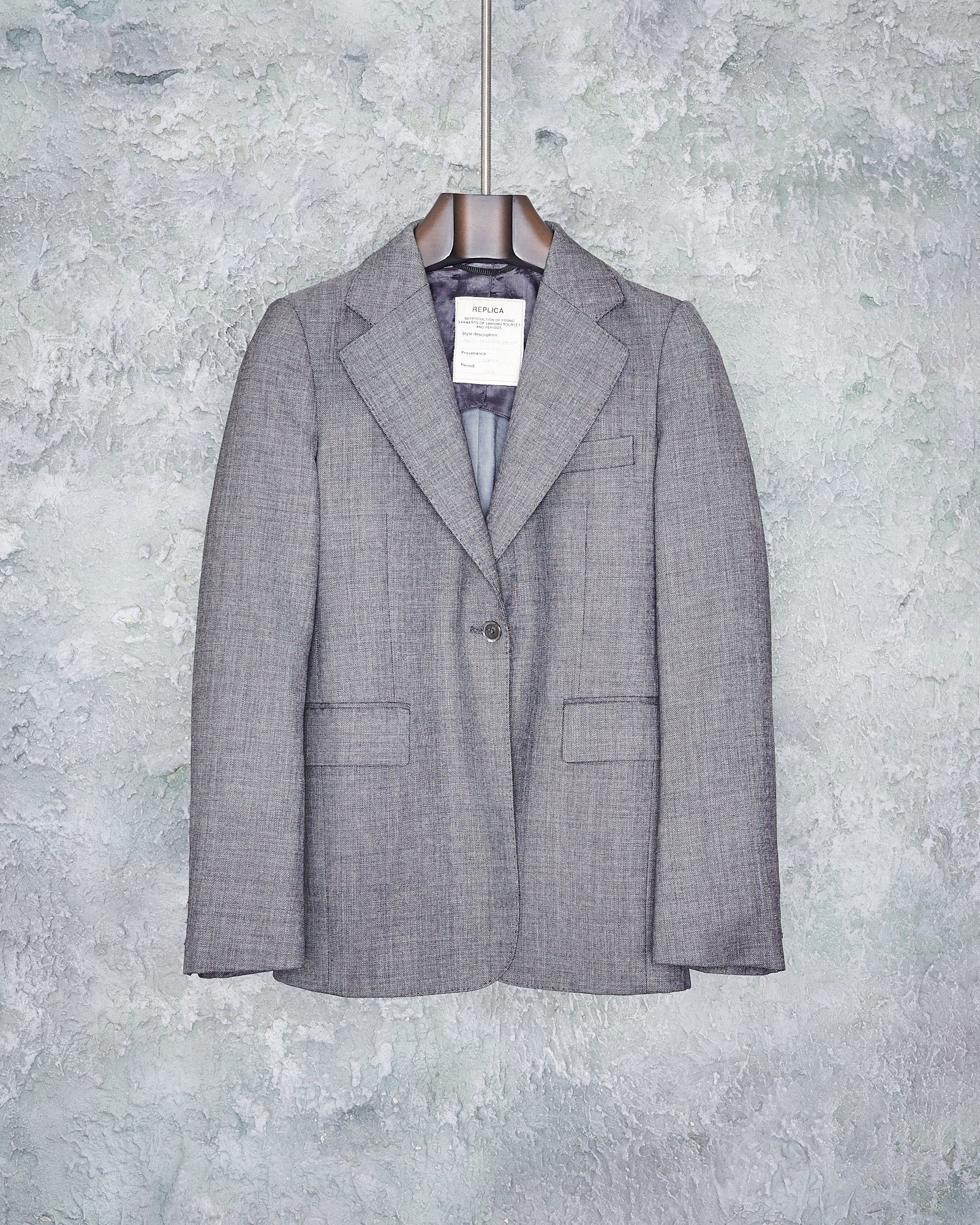 A gray wool Maison Margiela blazer with notch lapel and single button closure, displayed on a hanger against a textured background.
