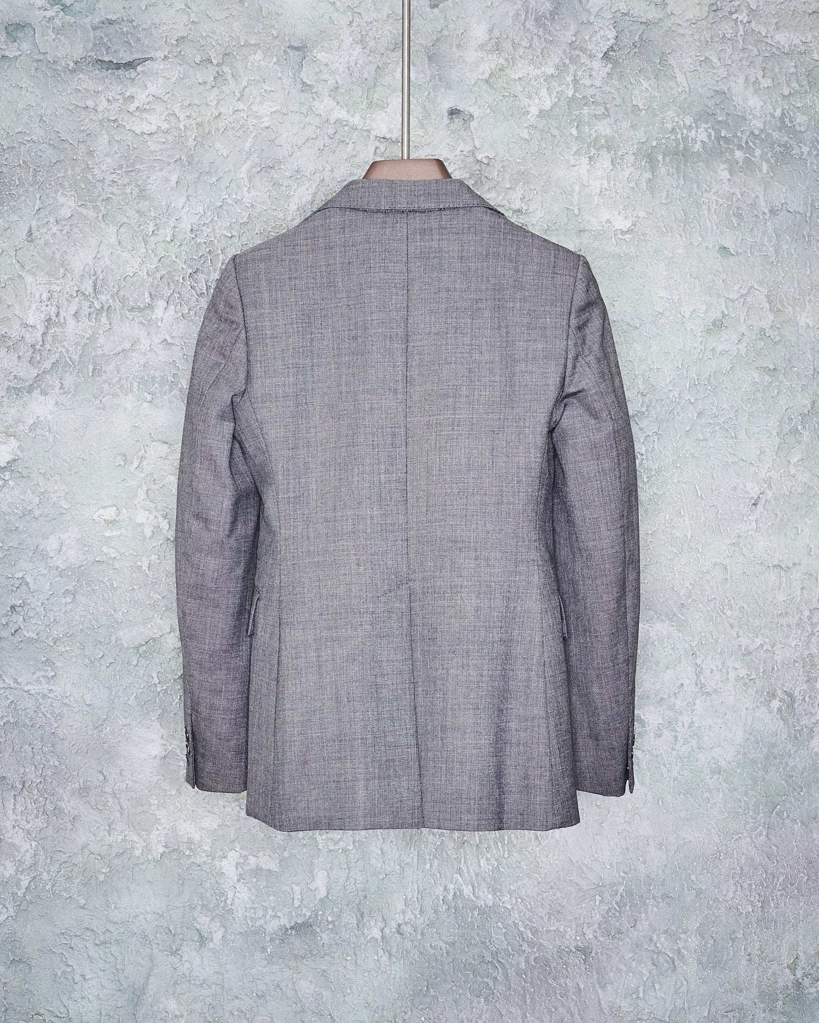 Reverse of a gray wool Maison Margiela blazer with notch lapel and single button closure, displayed on a hanger against a textured background.